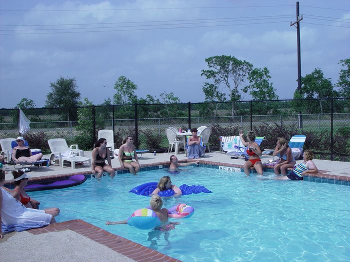 People at a pool party, some in the water with floats, others sitting around the pool, under a cloudy sky.