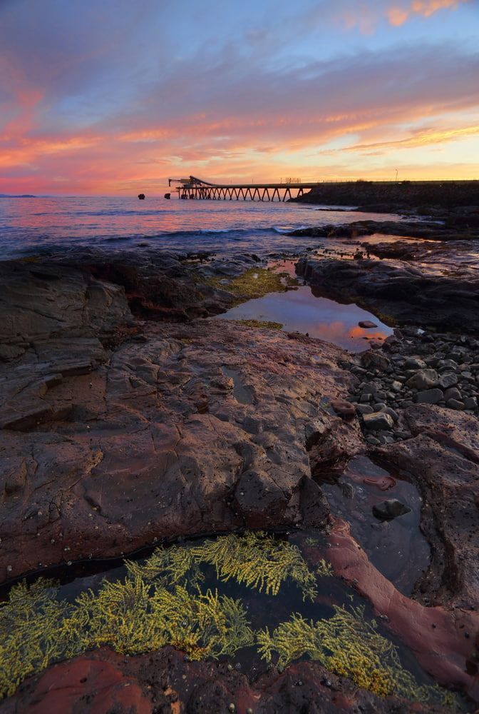 A Sunset Over a Rocky Beach with A Pier in The Distance — Leisure Coast Powder Coaters in Shellharbour, NSW