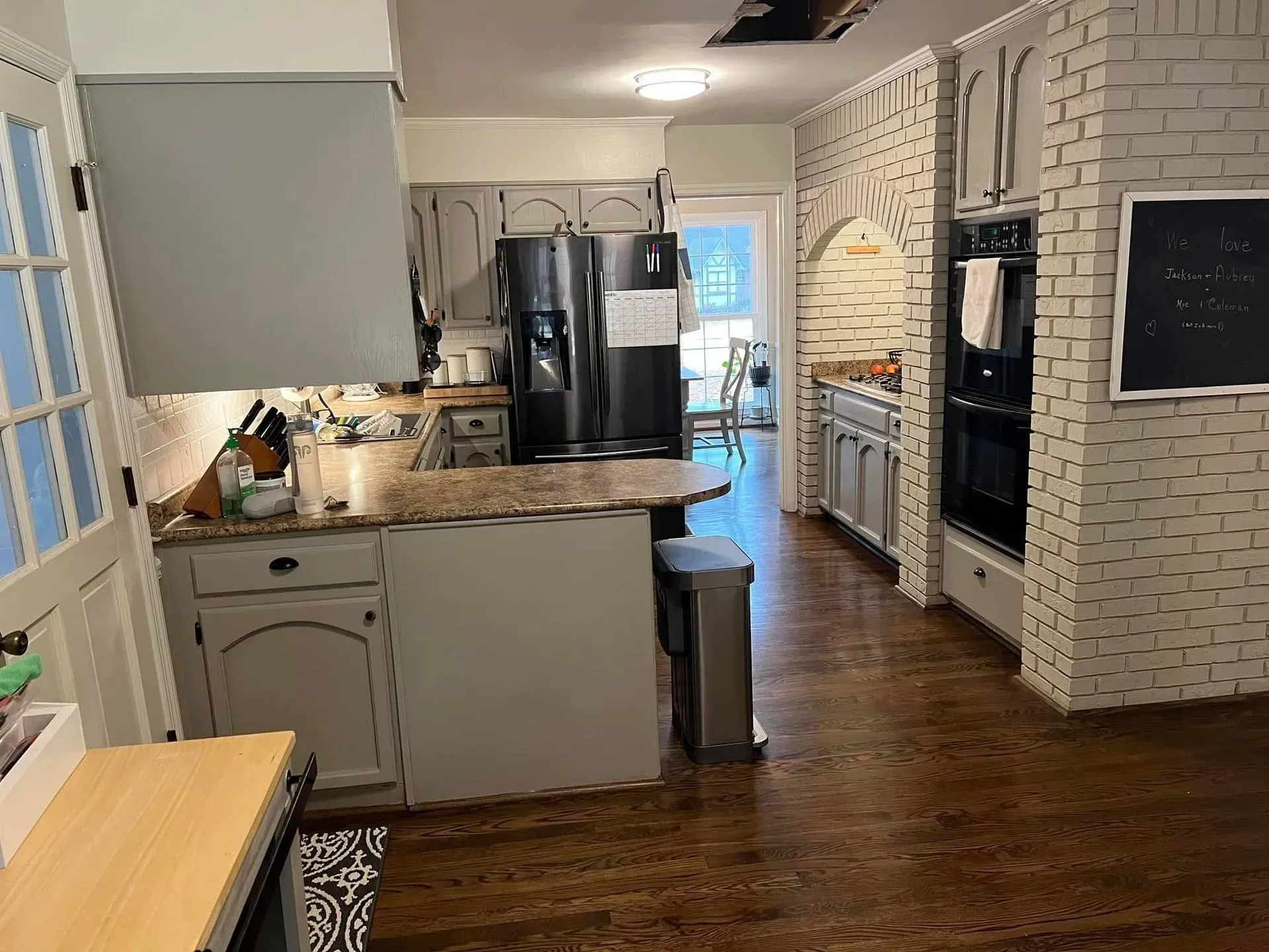 Kitchen with gray cabinets, light brick wall, and a dark refrigerator. Brown countertops and floors.