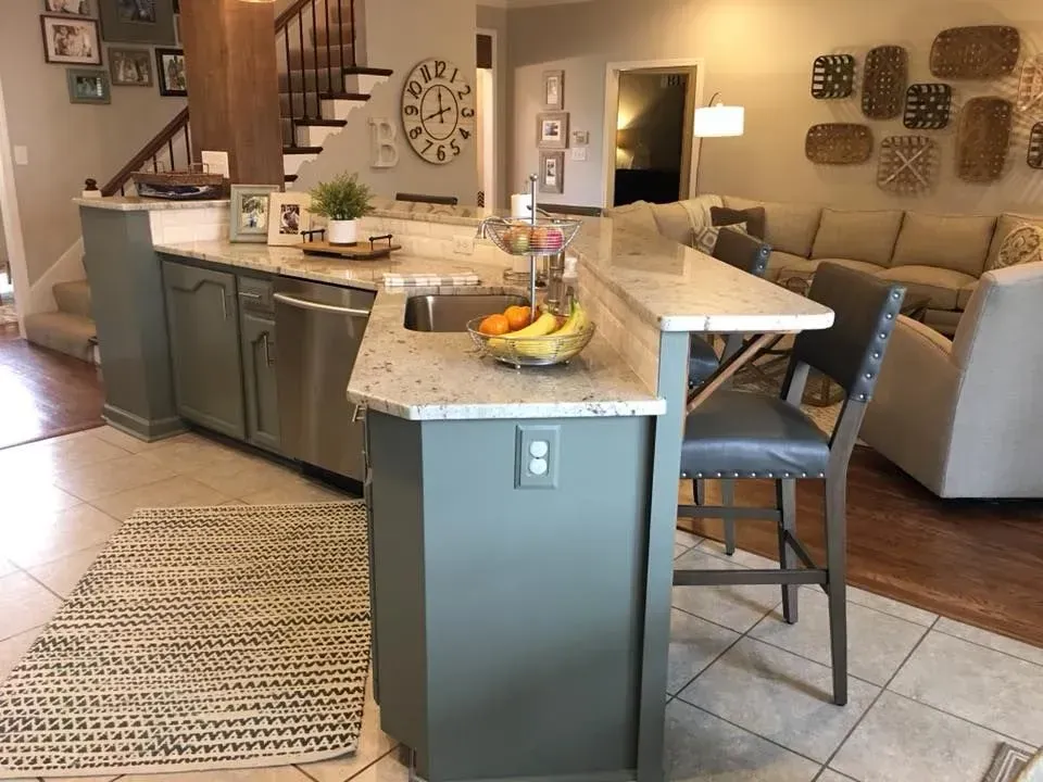 Kitchen island with gray-green cabinets and a granite countertop; includes a sink, bar stools, and a fruit bowl.