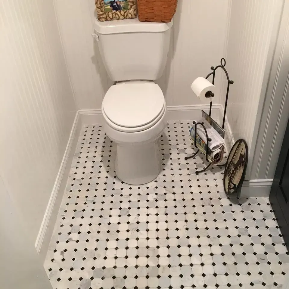A toilet in a small bathroom with black and white tile flooring. A decorative toilet paper stand is visible.