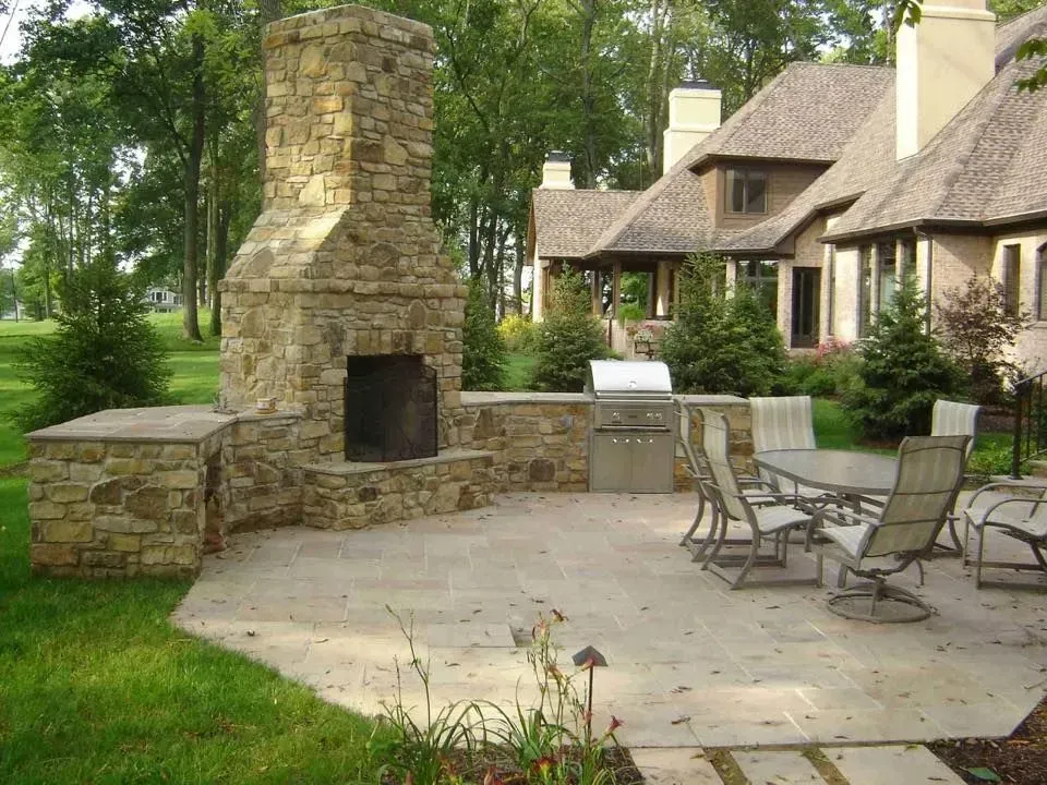 Stone outdoor fireplace and kitchen with a patio and seating area in a yard near a house.