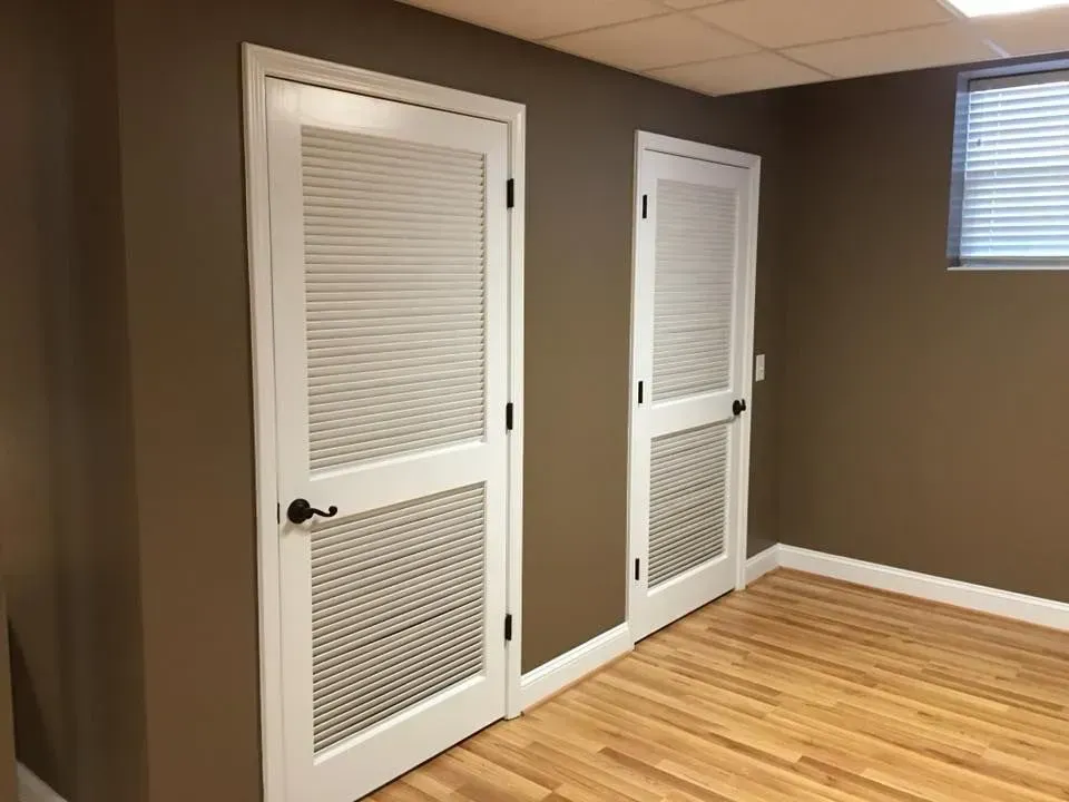 Two white, paneled doors with horizontal slats and a dark knob in a room with brown walls and wood flooring.