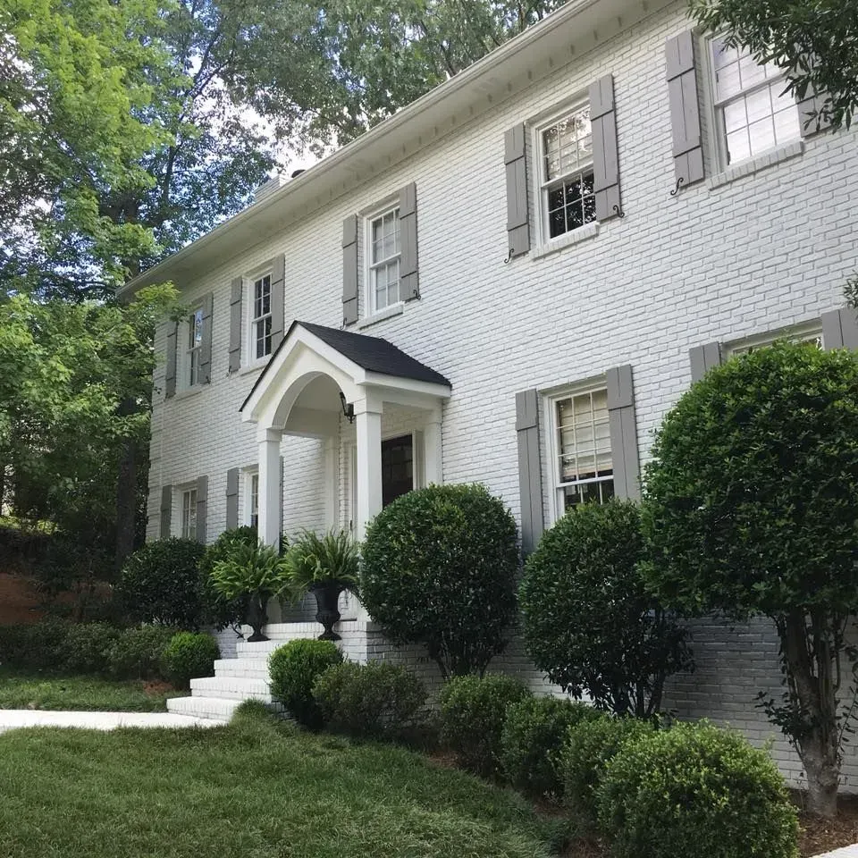Two-story white brick house with gray shutters and a small front porch. Green bushes and trees surround the home.