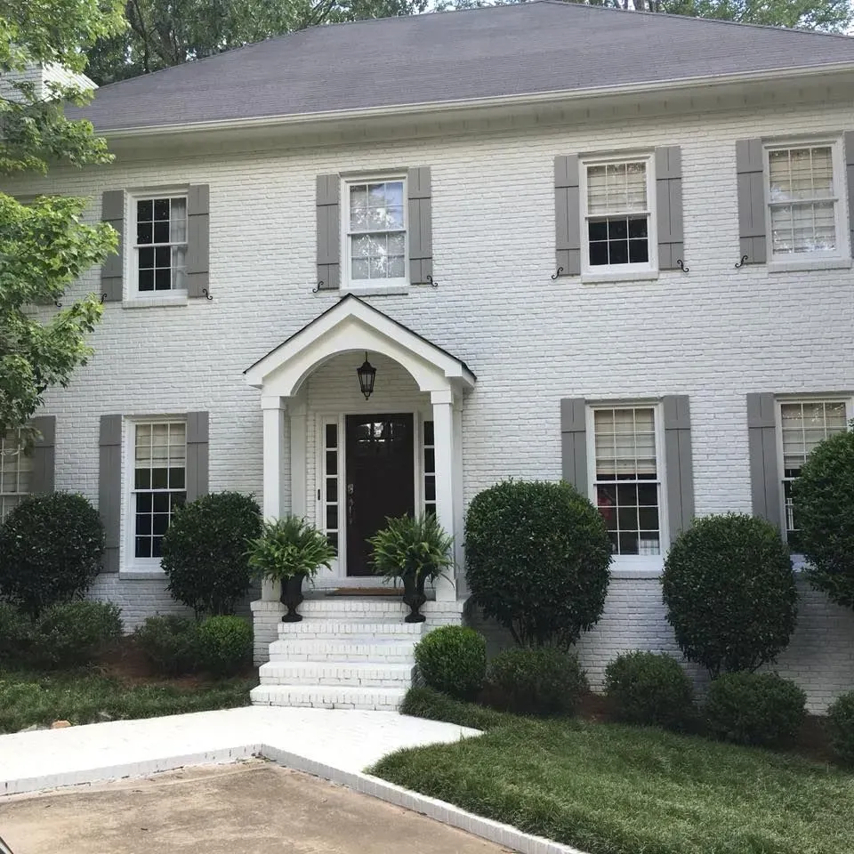 White brick two-story house with gray shutters and door. Landscaping includes bushes and a walkway.