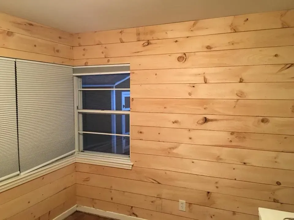 A room with wood plank walls surrounding a window with blinds. The wood is light-colored.
