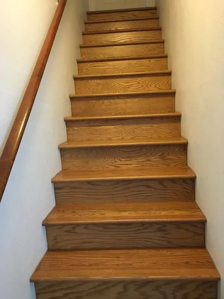 Wooden staircase with a handrail against a white wall, leading upwards.