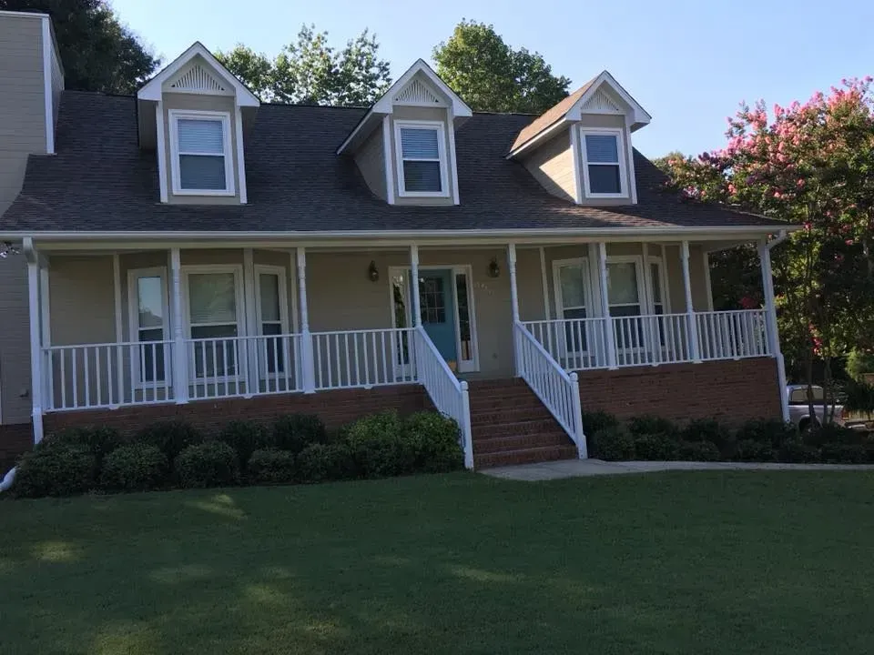 A light-colored house with a porch, brick foundation, and three dormer windows under a blue sky.