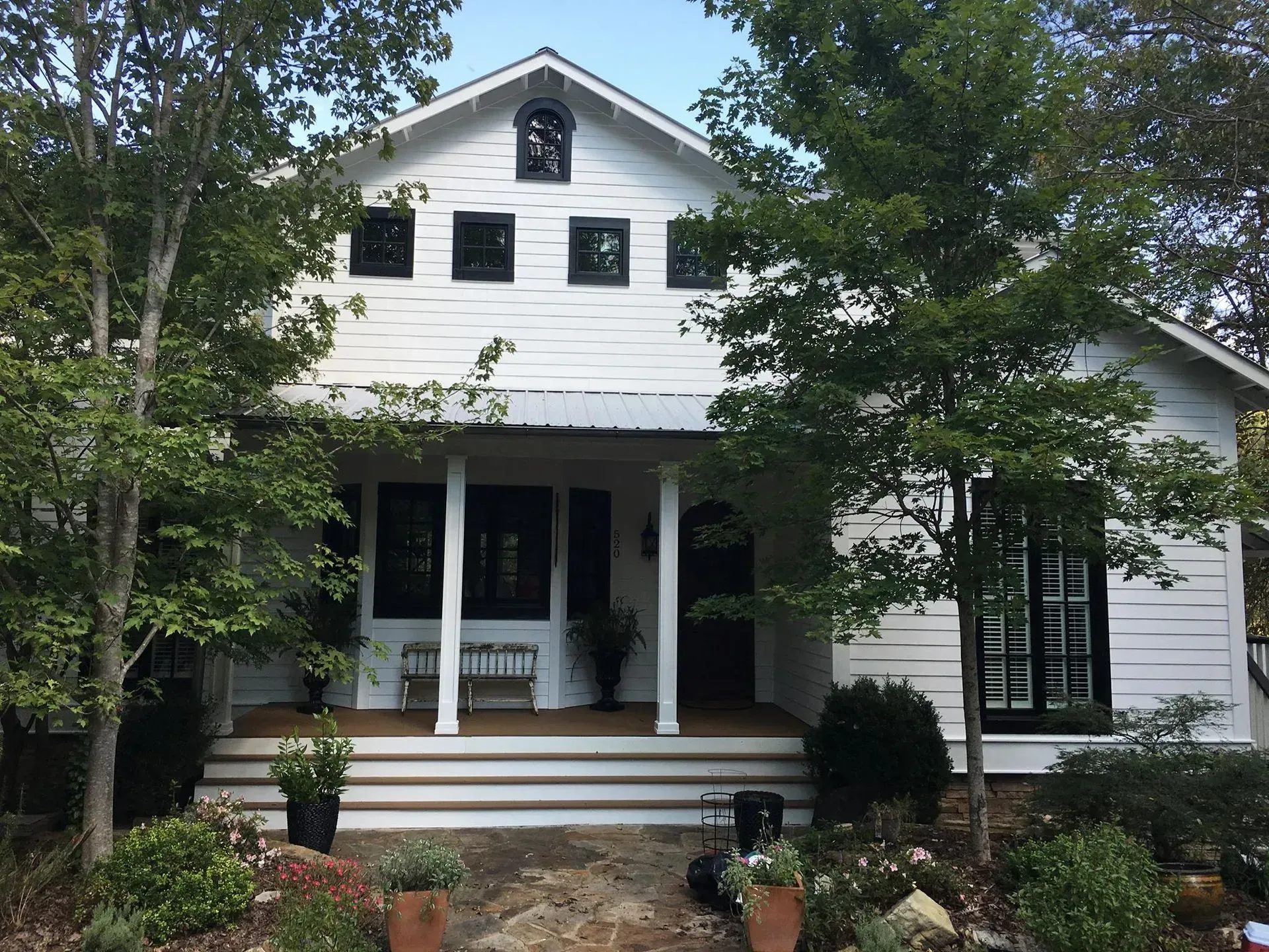 White farmhouse with black window frames and front door, a covered porch with white pillars, and landscaping.