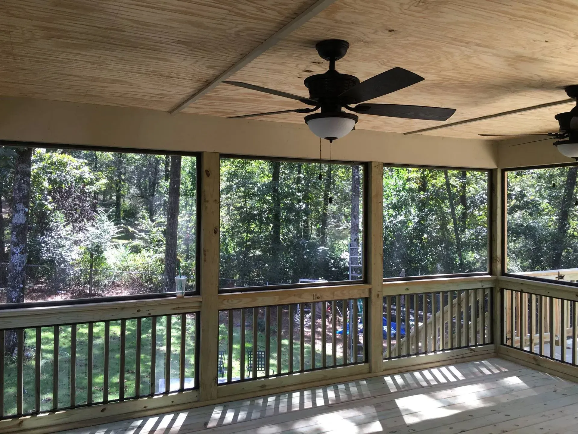 Screened-in porch with wooden railing and ceiling fans, overlooking a lush green forest.