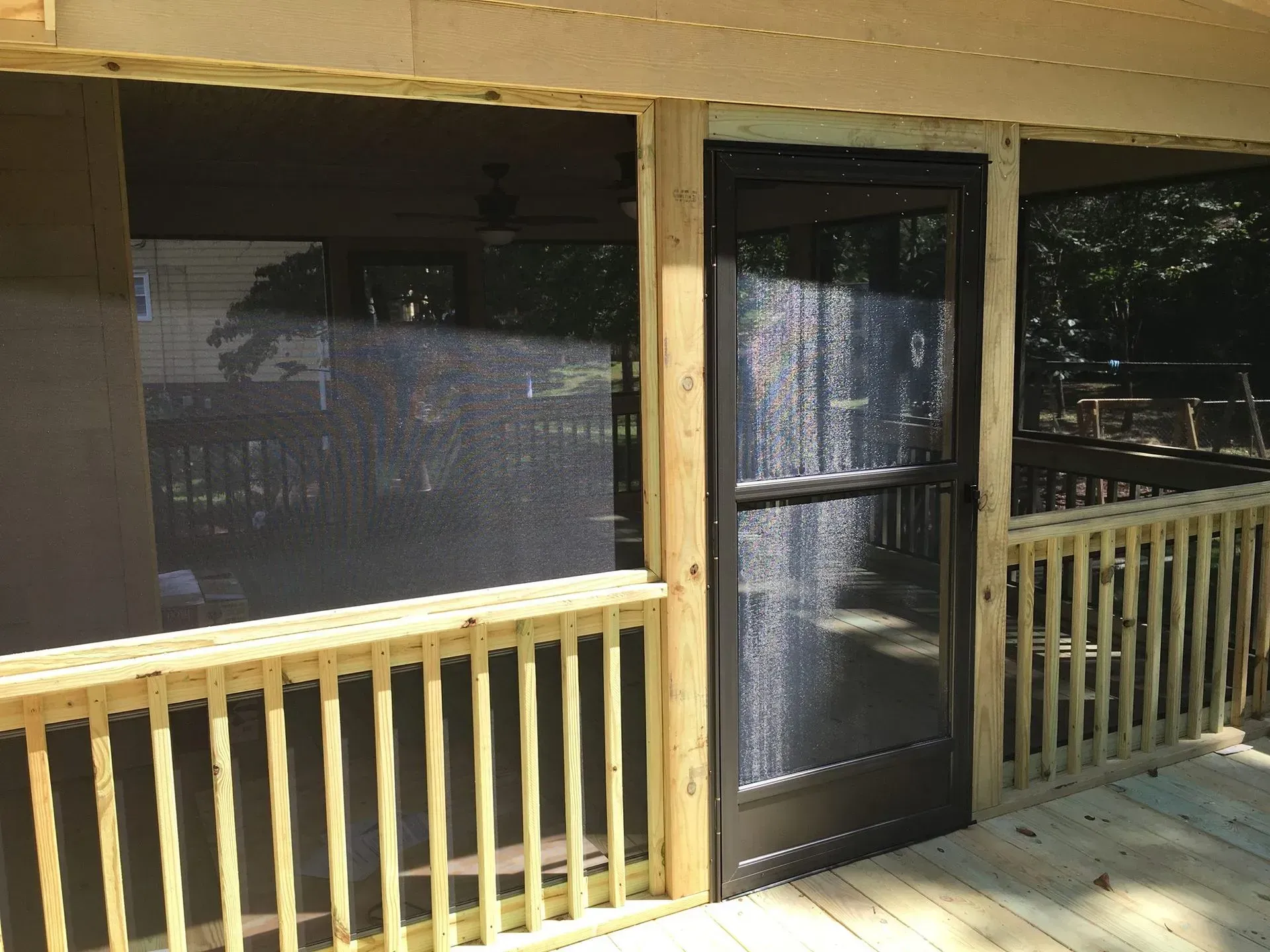 A screened porch with wooden railing and dark-framed screen door. Sunlight streams through the screens.