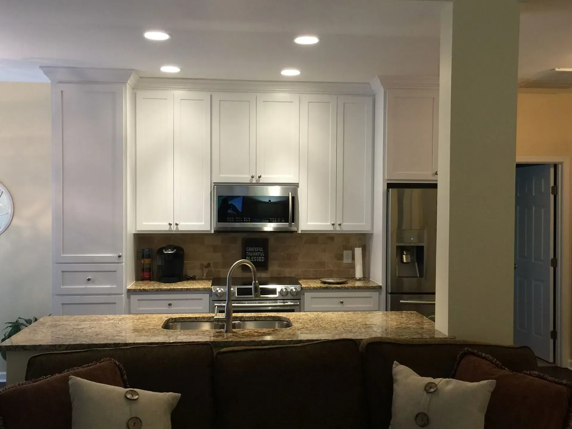 A kitchen with white cabinets, a stainless steel microwave, and a granite countertop. A brown sofa is in the foreground.