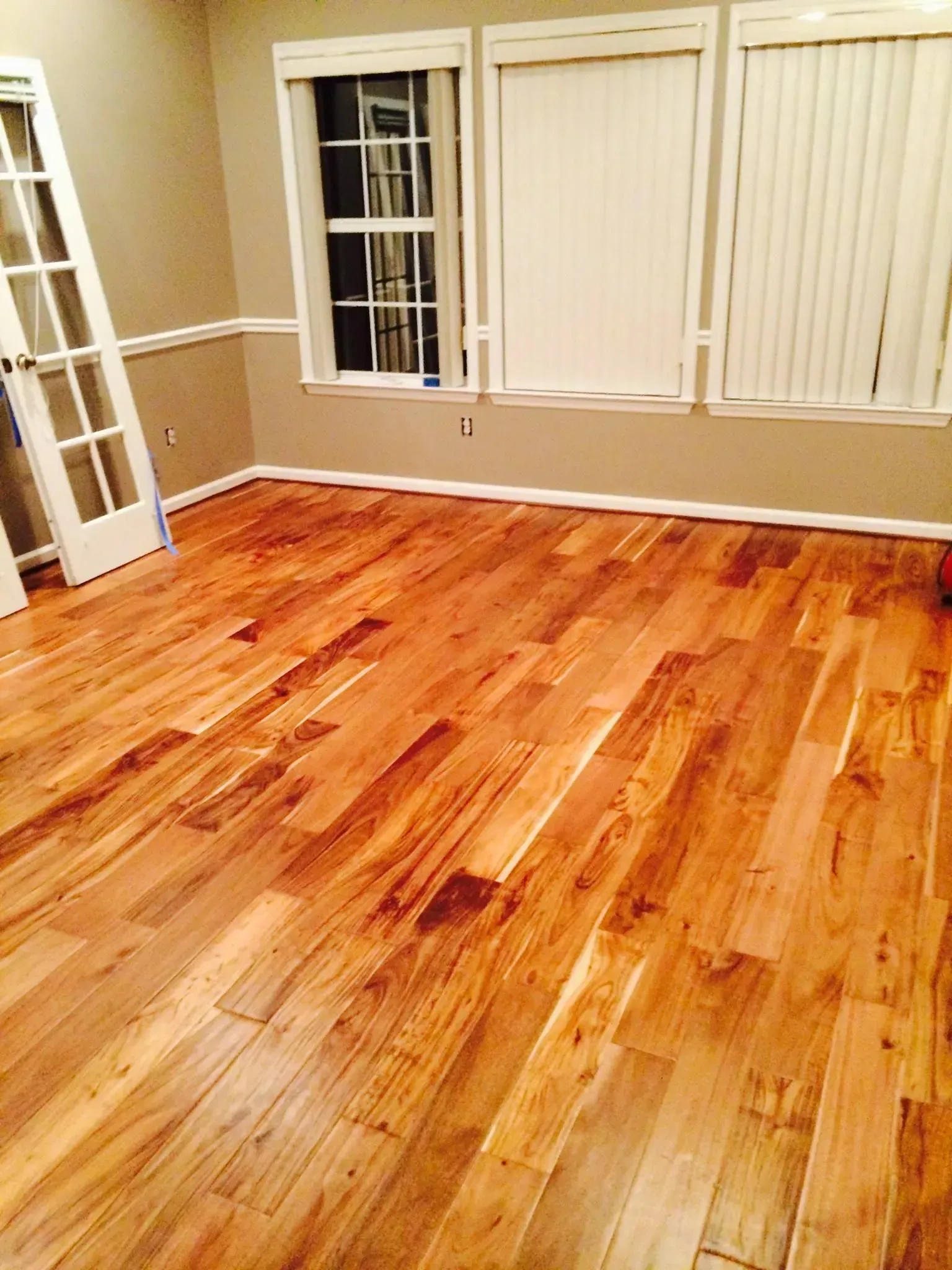 Hardwood flooring in a room with beige walls, white trim, and window blinds. A door is partially visible on the left.