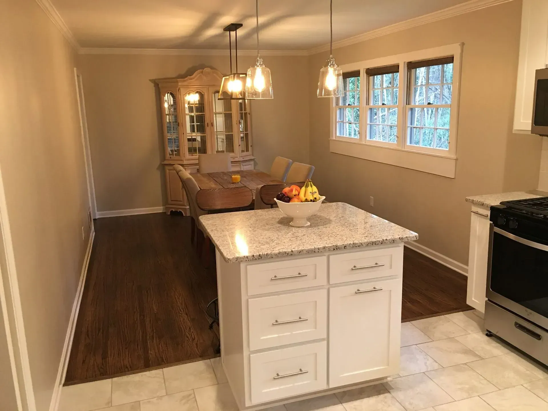 A kitchen with a white island, wooden floors, dining area with a china cabinet, and pendant lights.