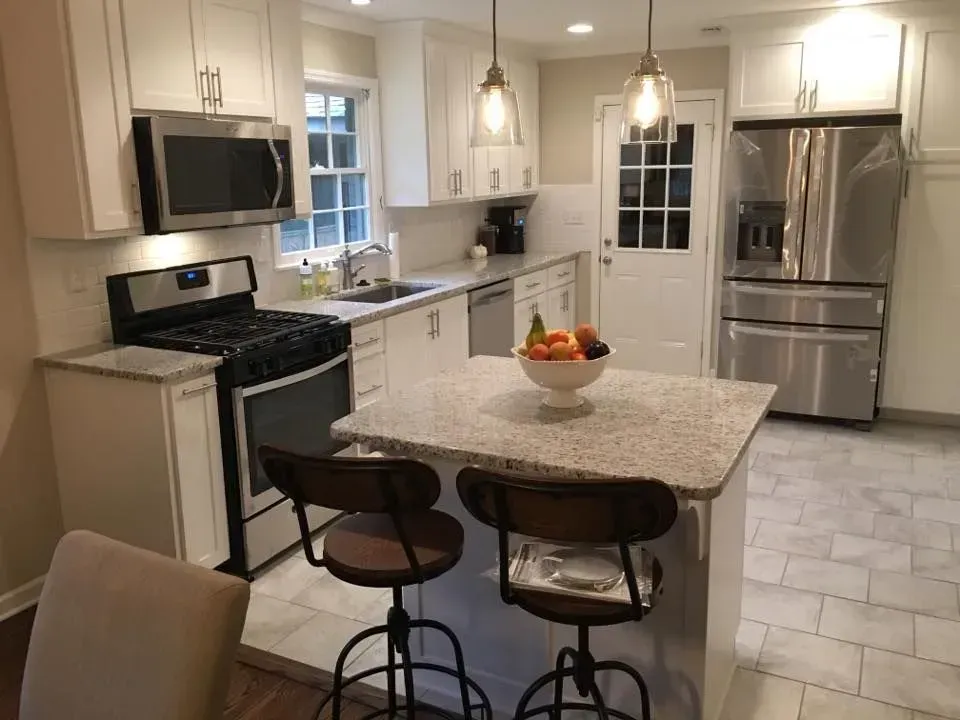 A bright, renovated kitchen with white cabinets, stainless steel appliances, and a small island with two stools.