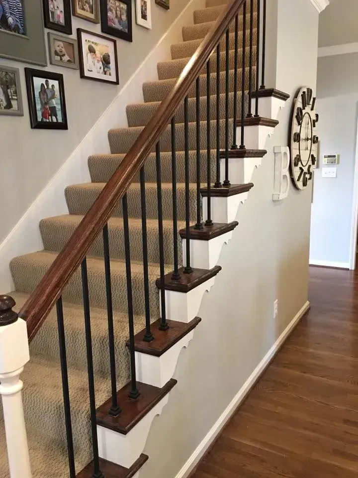 Staircase with brown handrail and carpeted steps, black metal spindles, white trim, and wooden flooring in a home.