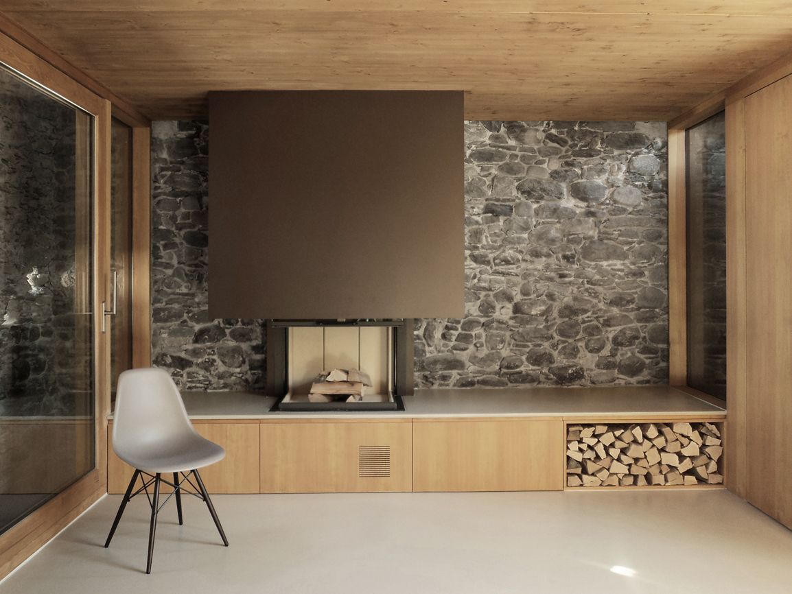 Stanza moderna con camino in pietra, pannelli in legno e una sedia grigia. Deposito per la legna da ardere sottostante.
