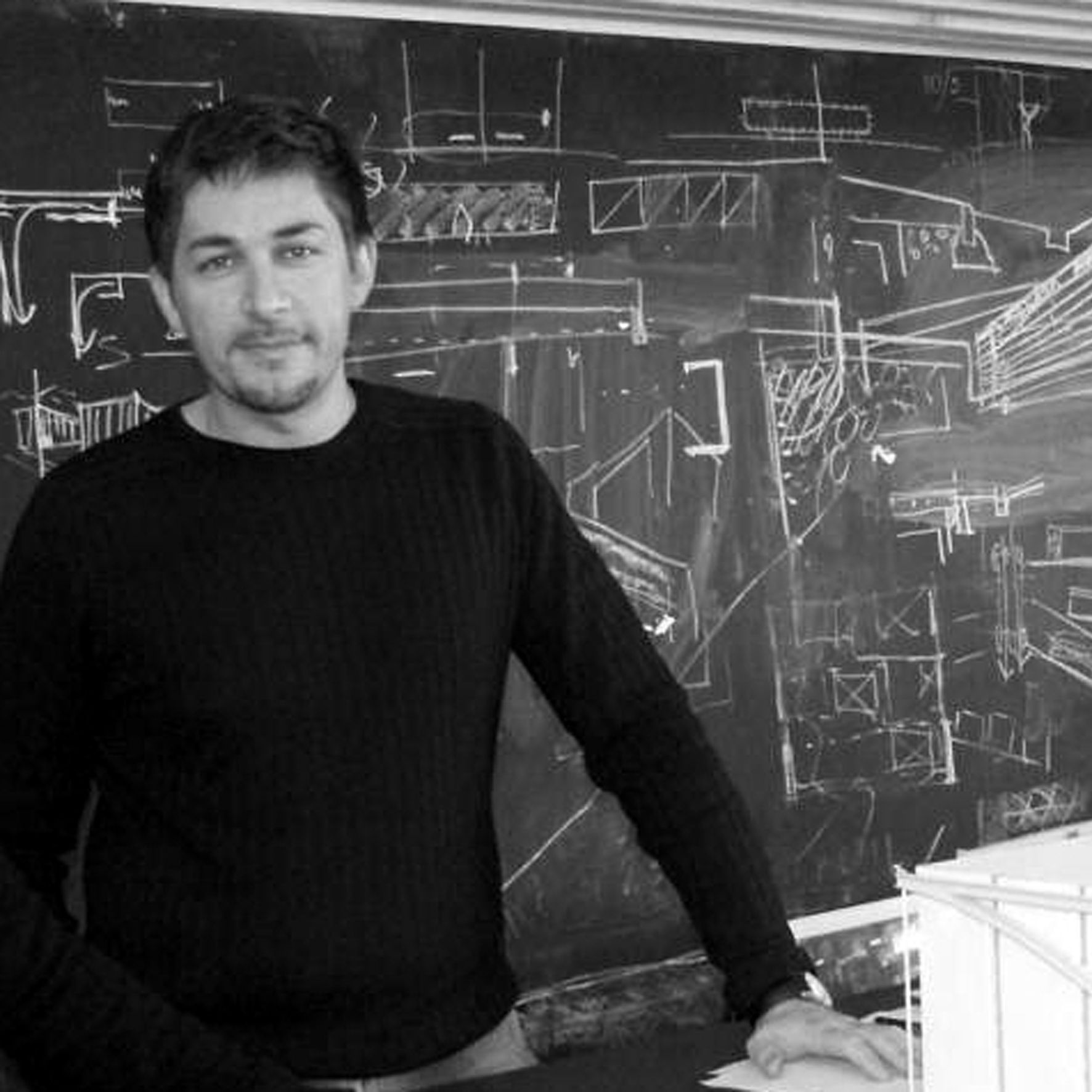 Un uomo in piedi davanti a una lavagna con schizzi architettonici. Indossa un maglione nero e sorride leggermente.