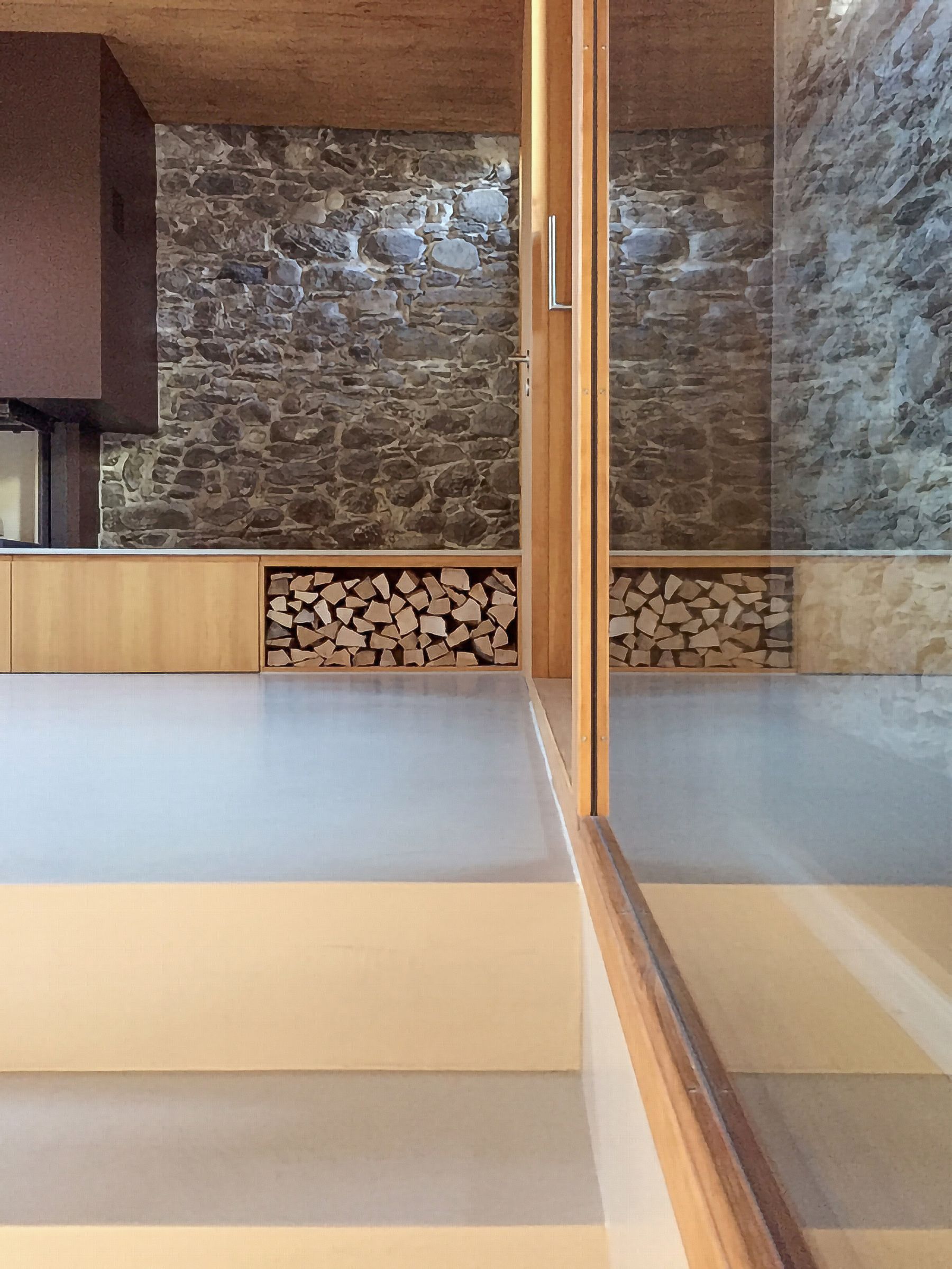 Interno con parete in pietra, legnaia e porta in vetro che riflette la stanza.
