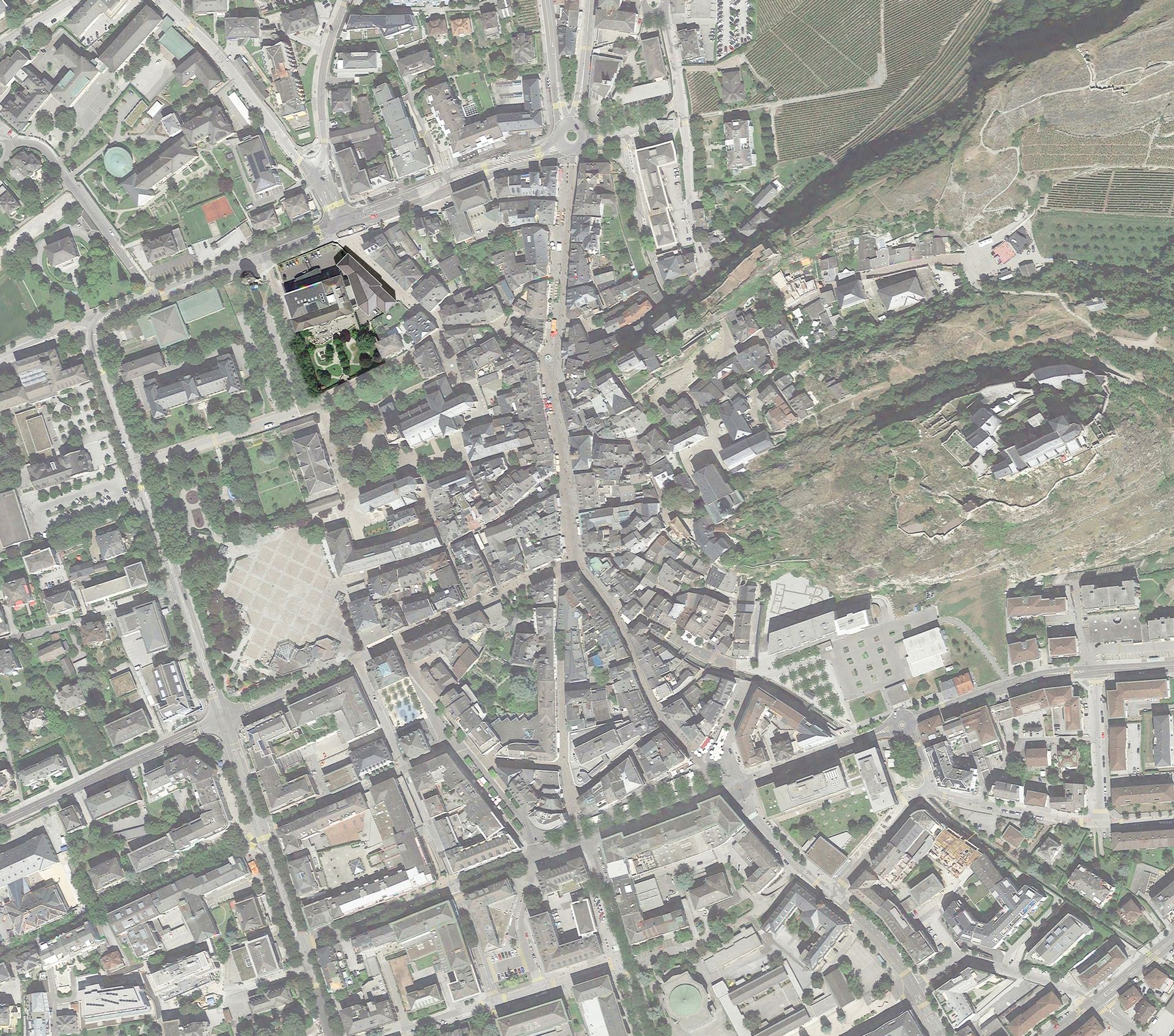 Vista aerea di una città con una strada centrale, edifici, spazi verdi e una collina.