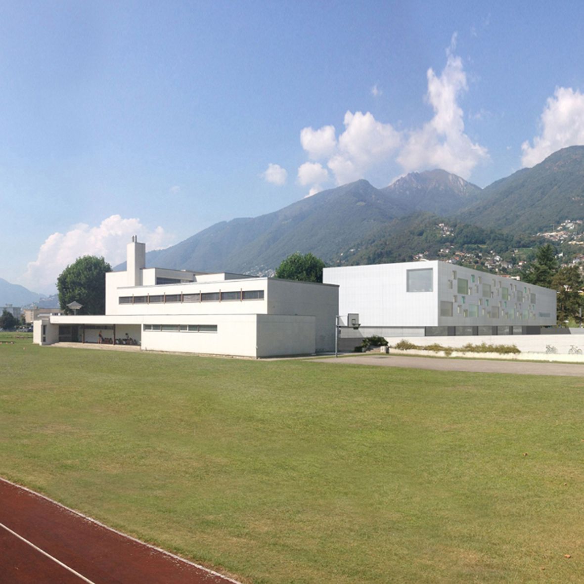 Edifici bianchi moderni con un campo erboso e montagne sullo sfondo sotto un cielo azzurro.