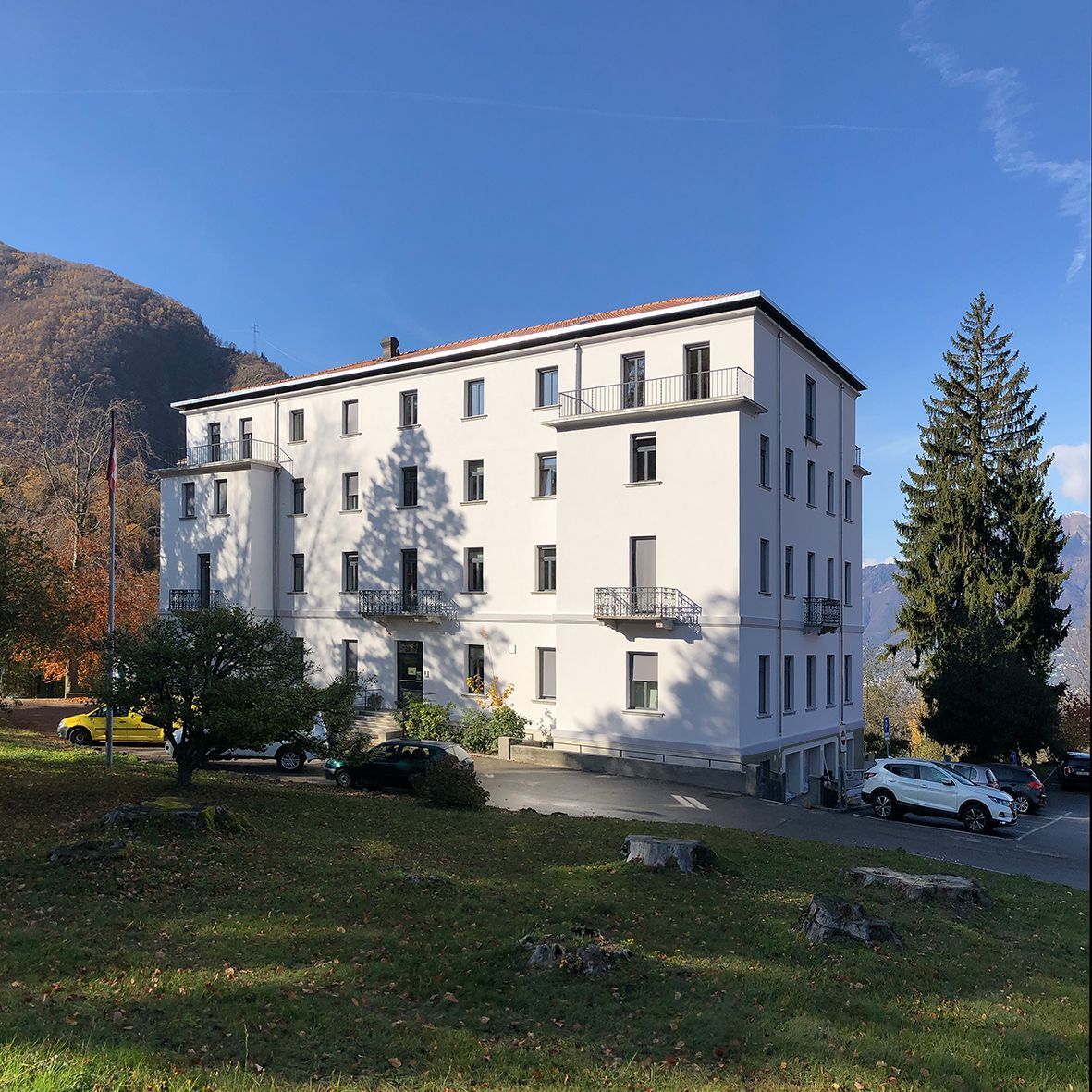 Edificio bianco con numerose finestre e balconi in una giornata di sole, con alberi e una montagna sullo sfondo.