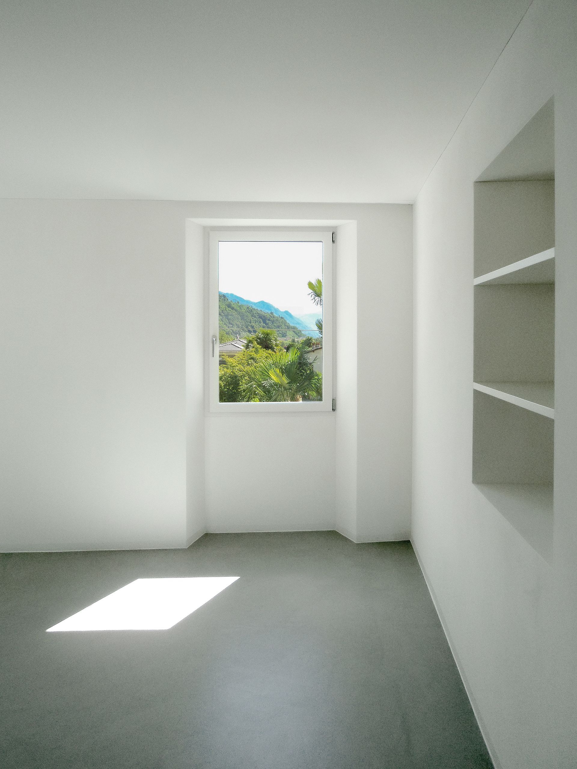 Stanza bianca e vuota con finestra che si affaccia su un paesaggio montano; scaffalature integrate sulla destra.