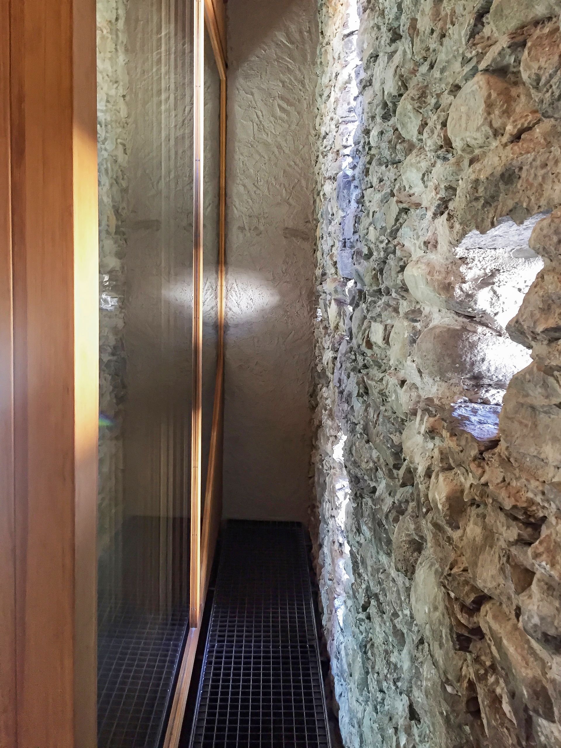 Corridoio stretto con pareti in pietra, pavimento in griglia metallica, luce solare intensa e porta in legno.