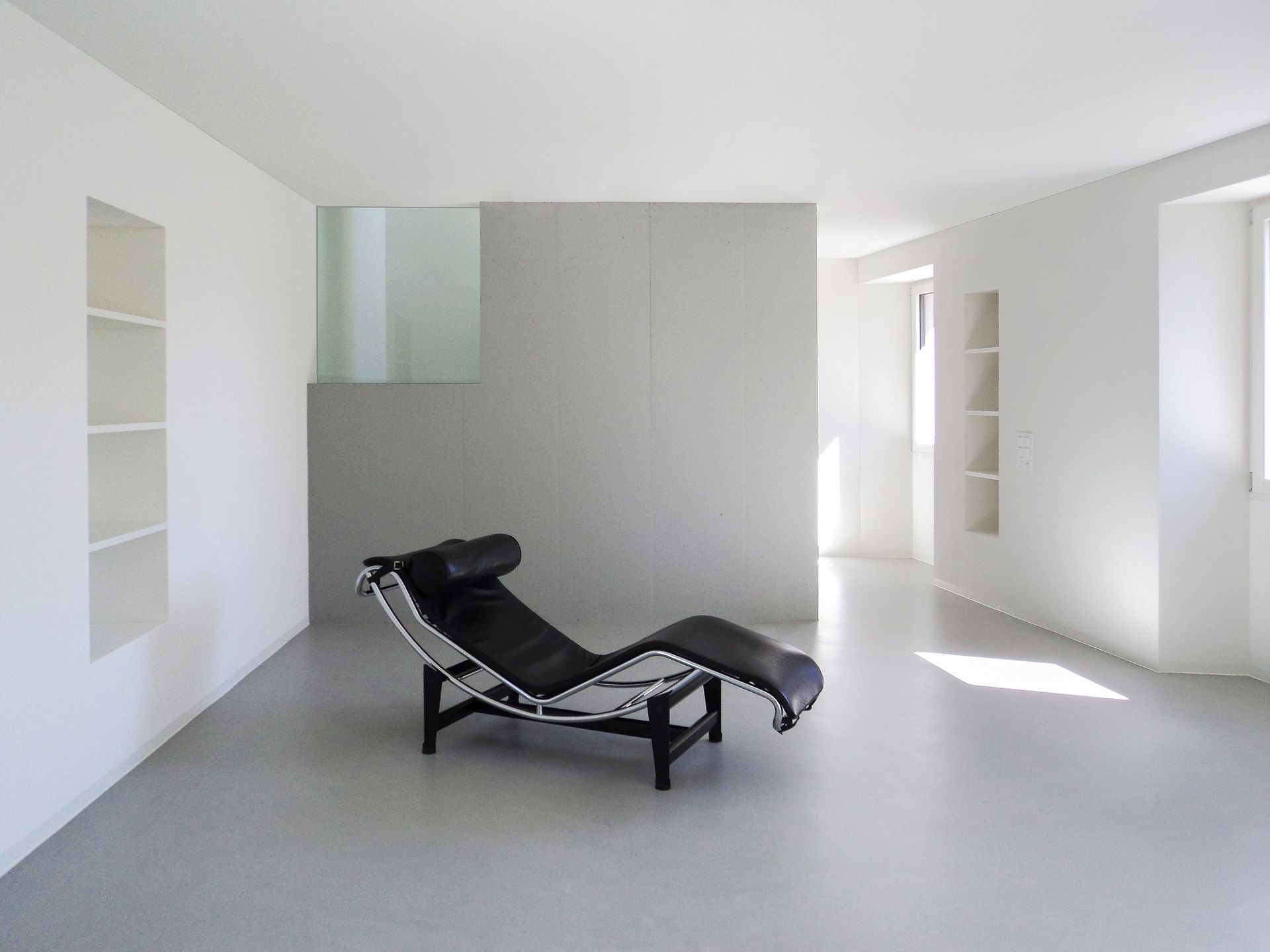 Stanza bianca minimalista con chaise longue nera, mensole incassate e una parete di cemento.