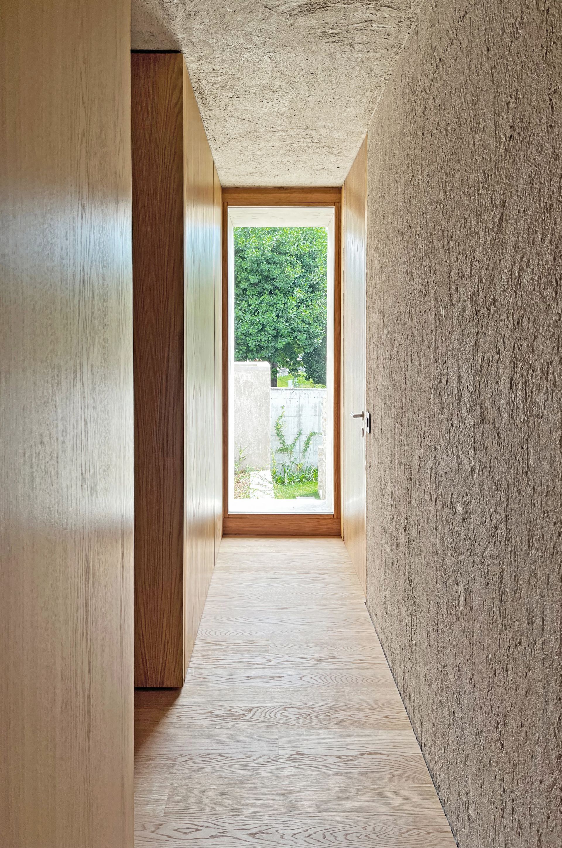 Corridoio stretto con pareti e soffitto in intonaco grezzo e legno che conduce a una luminosa vista esterna.