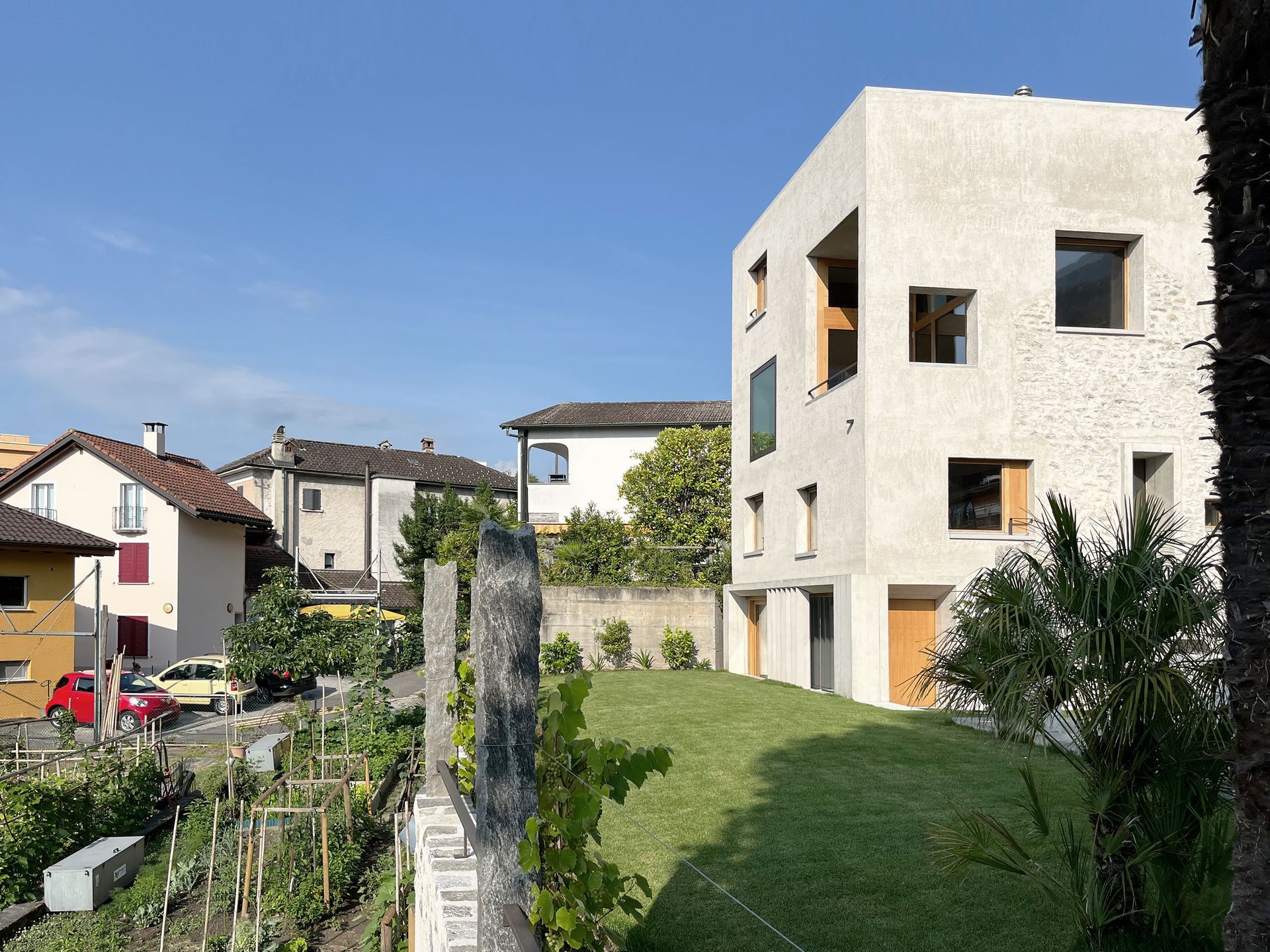 Moderno edificio in pietra e cemento con finestre quadrate, altri edifici sullo sfondo, prato verde, cielo azzurro.