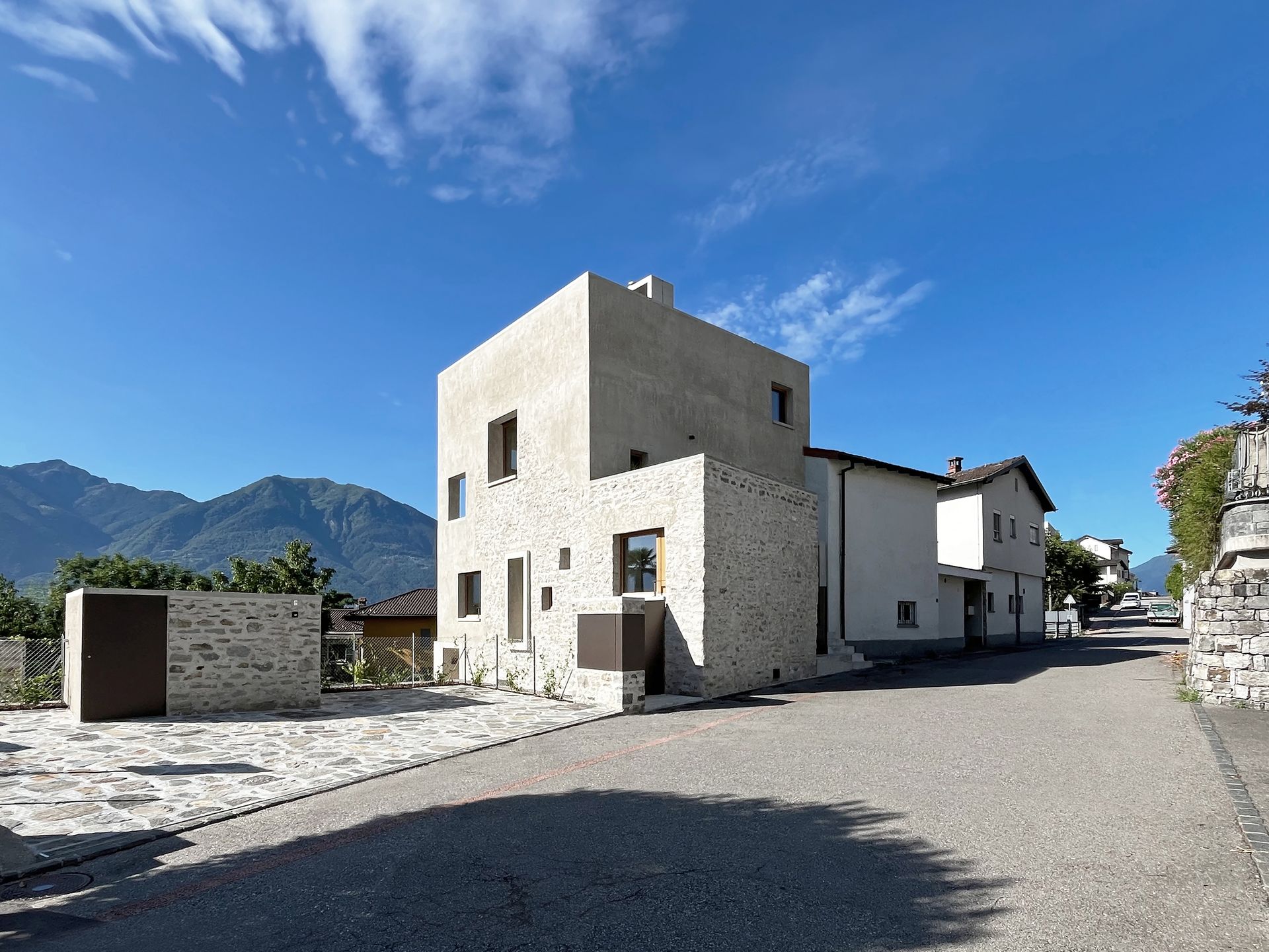 Moderno edificio in pietra e cemento sotto un cielo azzurro, strada in primo piano, montagne sullo sfondo.