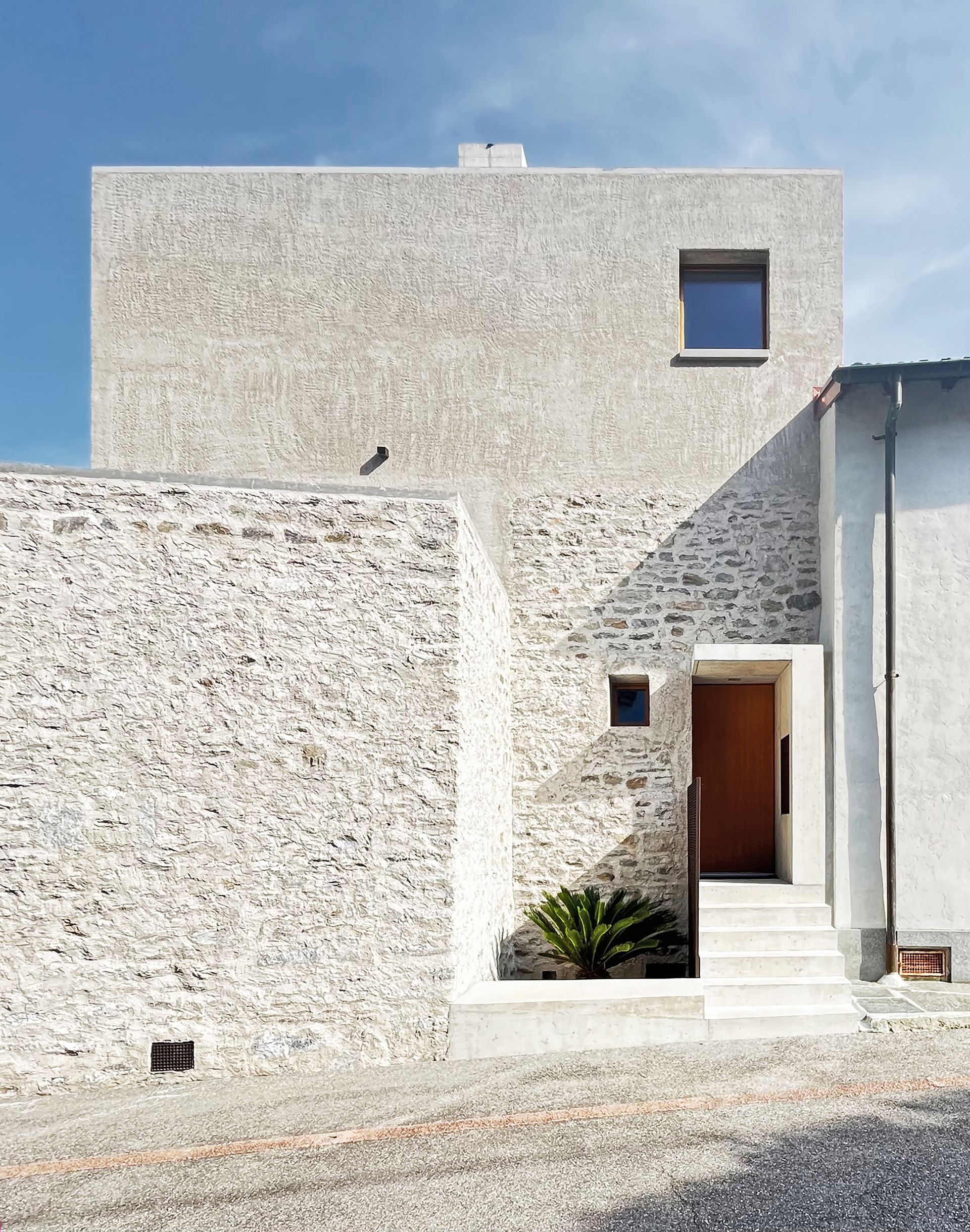 Casa moderna con pareti esterne in rasa pietra, colori chiari e un piccolo muro che circonda l'ingresso.