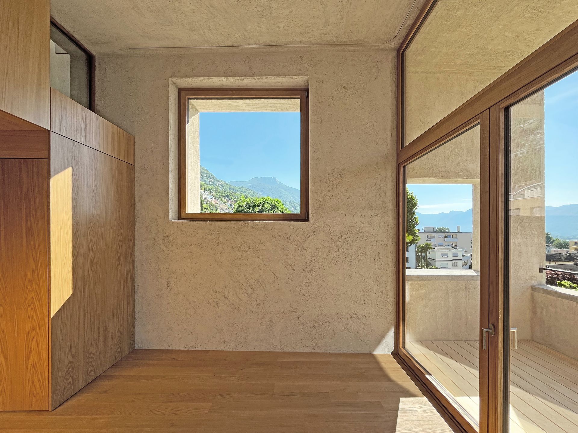 Vista interna di una stanza con pavimenti in legno, pareti strutturate, una vetrata a destra che conduce a un balcone.