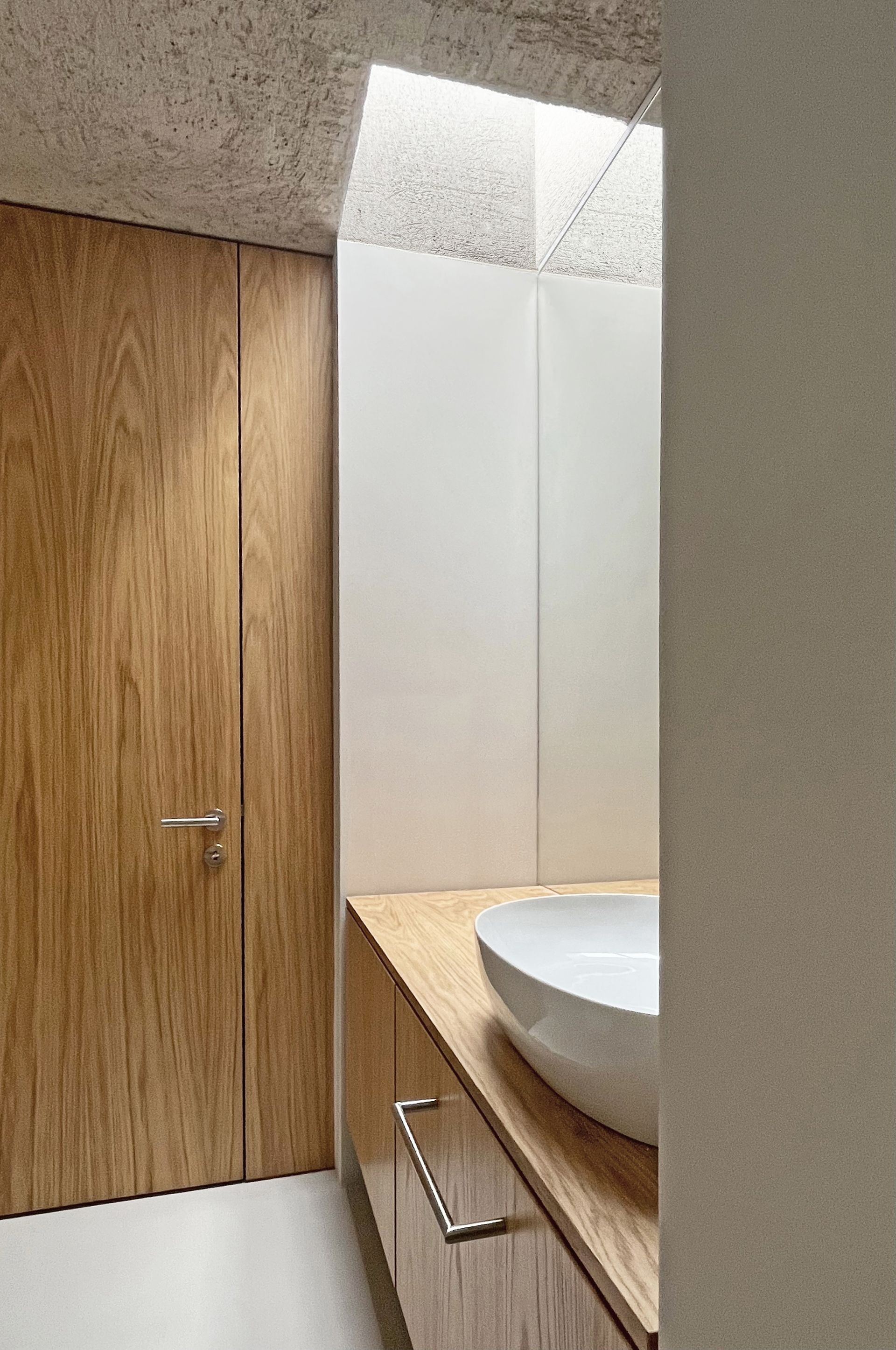 Bagno con porta in legno, mobile lavabo e lavandino bianco. La luce filtra dall'alto.