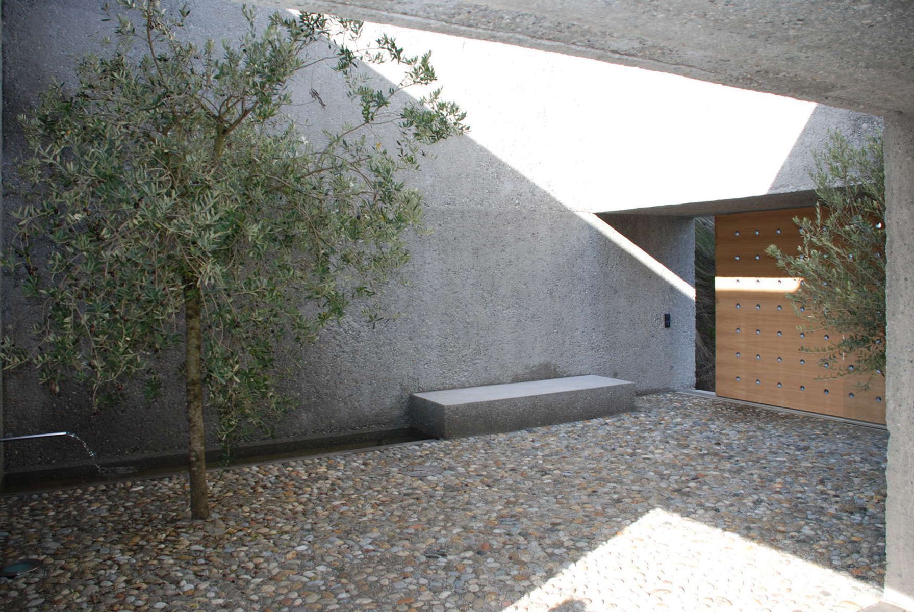 Cortile con ulivi, terreno ghiaioso, muri di cemento e una panchina in pietra.