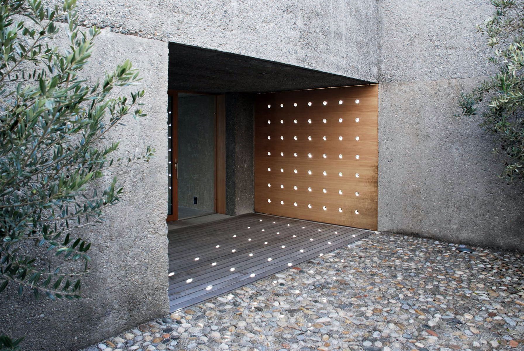 Ingresso con rivestimento in cemento strutturato. Sono visibili un pannello in legno con fori e un sentiero in ghiaia.