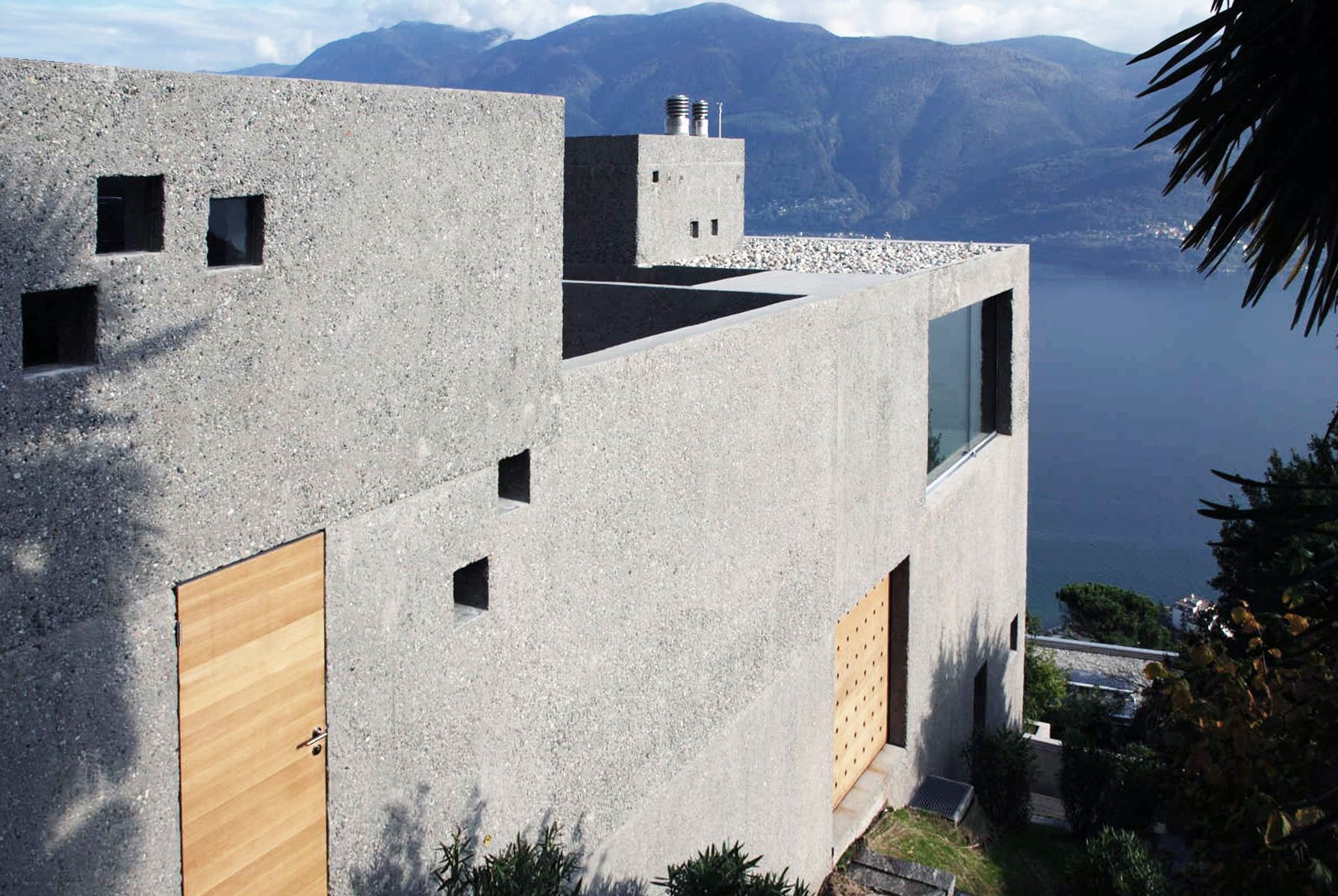 Edificio in cemento con porte in legno e finestre quadrate, con vista sul lago e sulle montagne.
