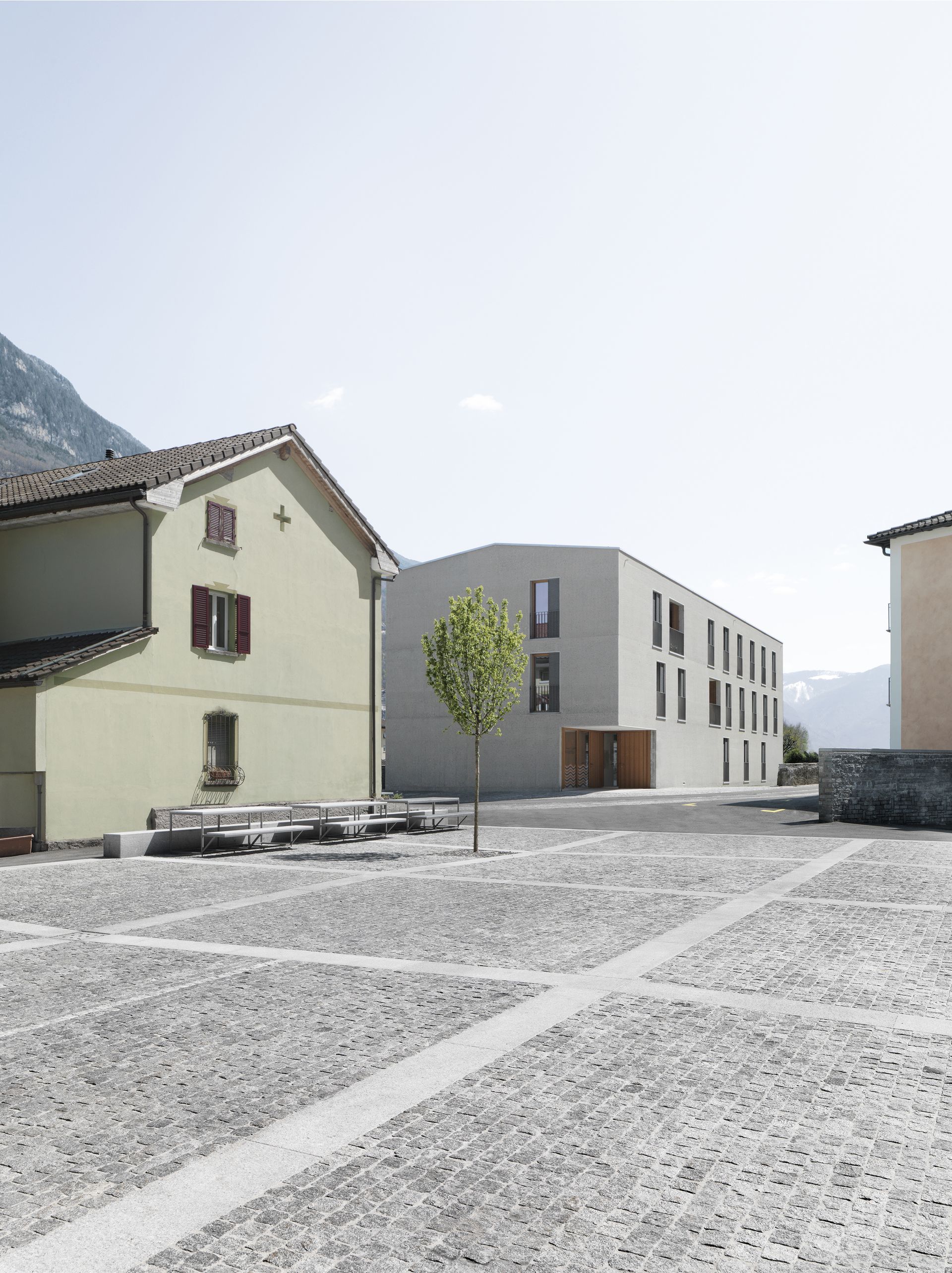 Un edificio moderno e una casa più antica si trovano su una piazza acciottolata. Tra di loro c'è un piccolo albero.