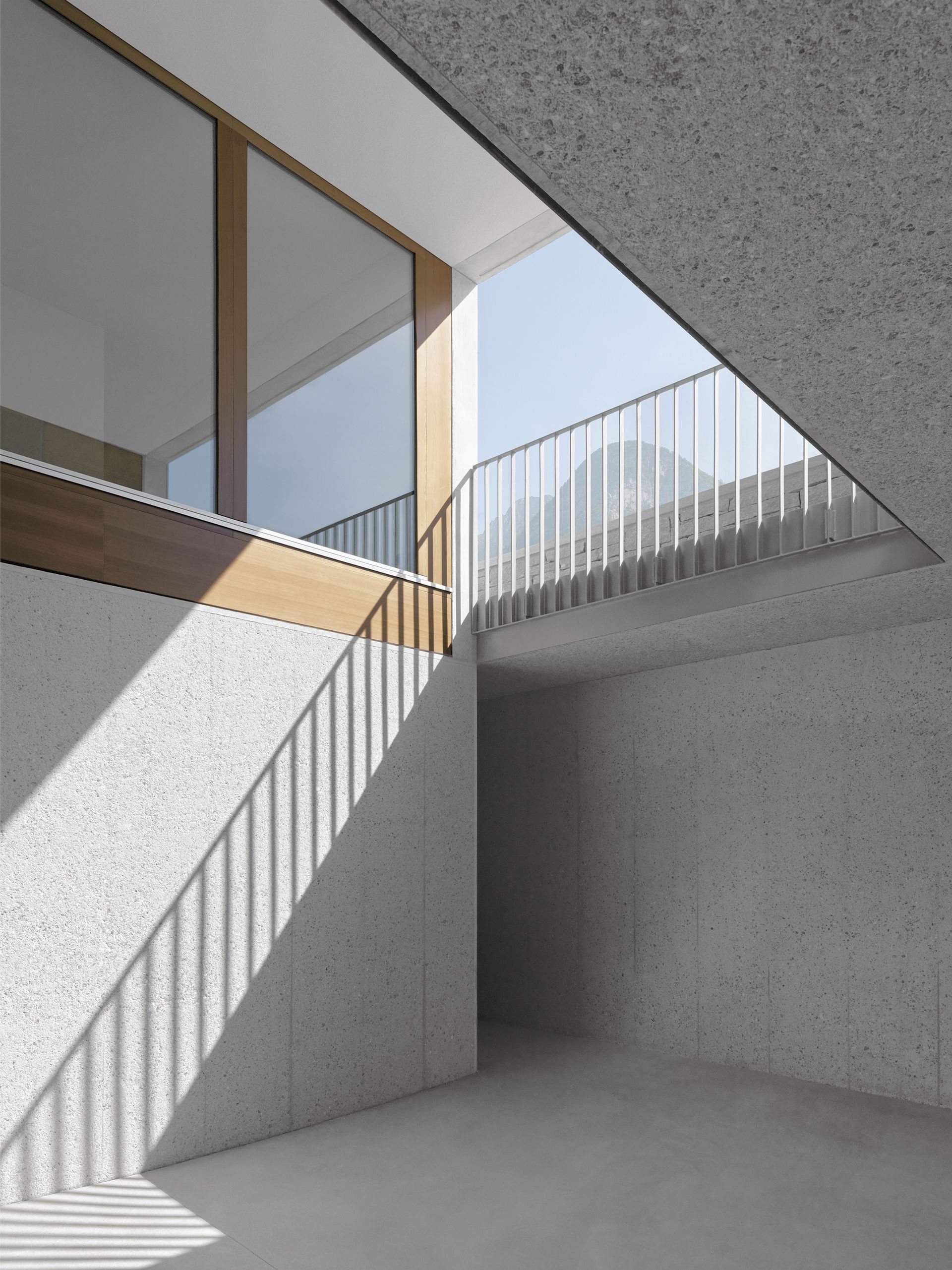 Interni dall'architettura moderna. Pareti in cemento illuminate dal sole, infissi in legno, balcone con ringhiera in metallo.