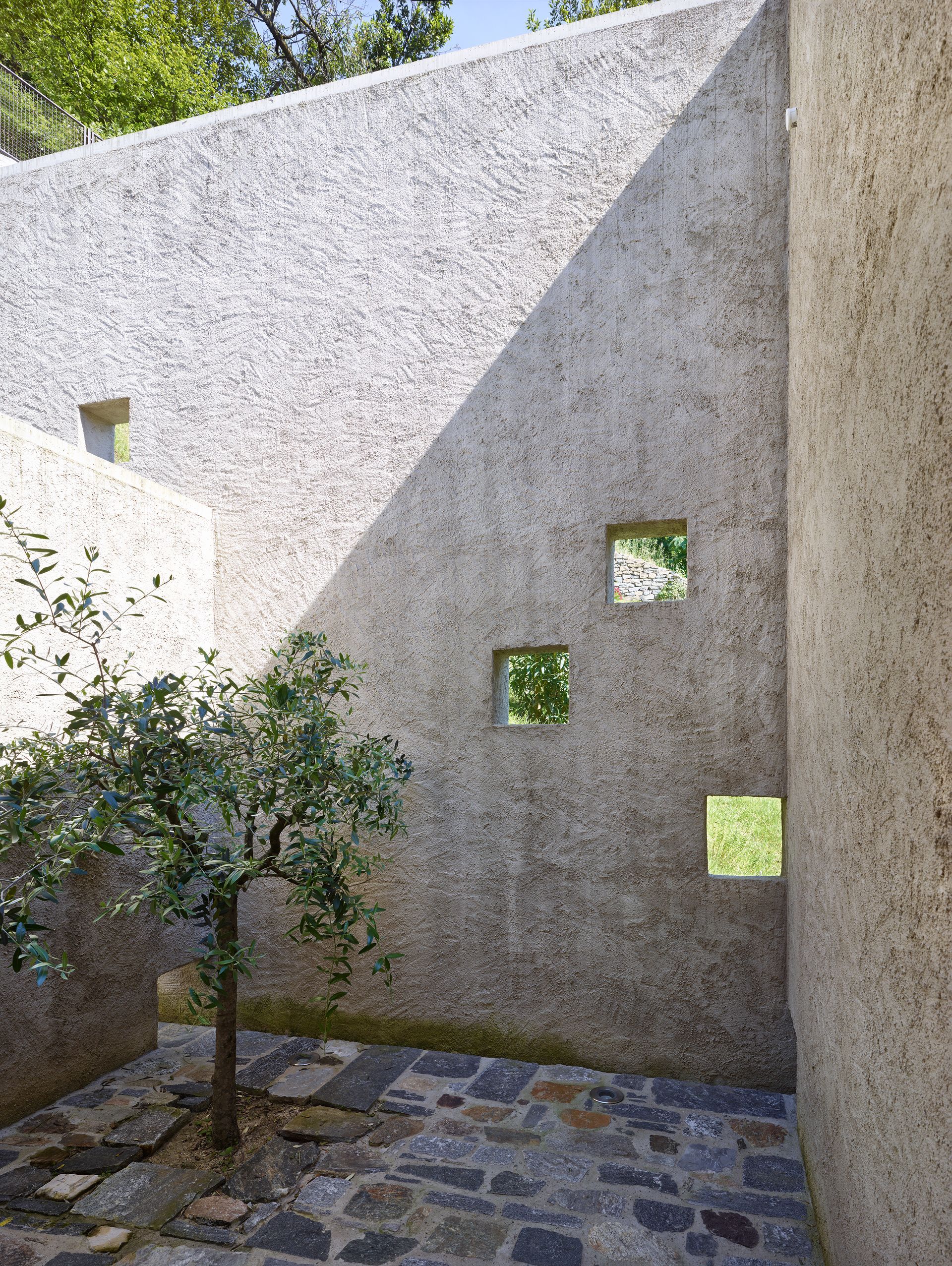 Cortile in pietra con alberi, luce e aperture quadrate nei muri.