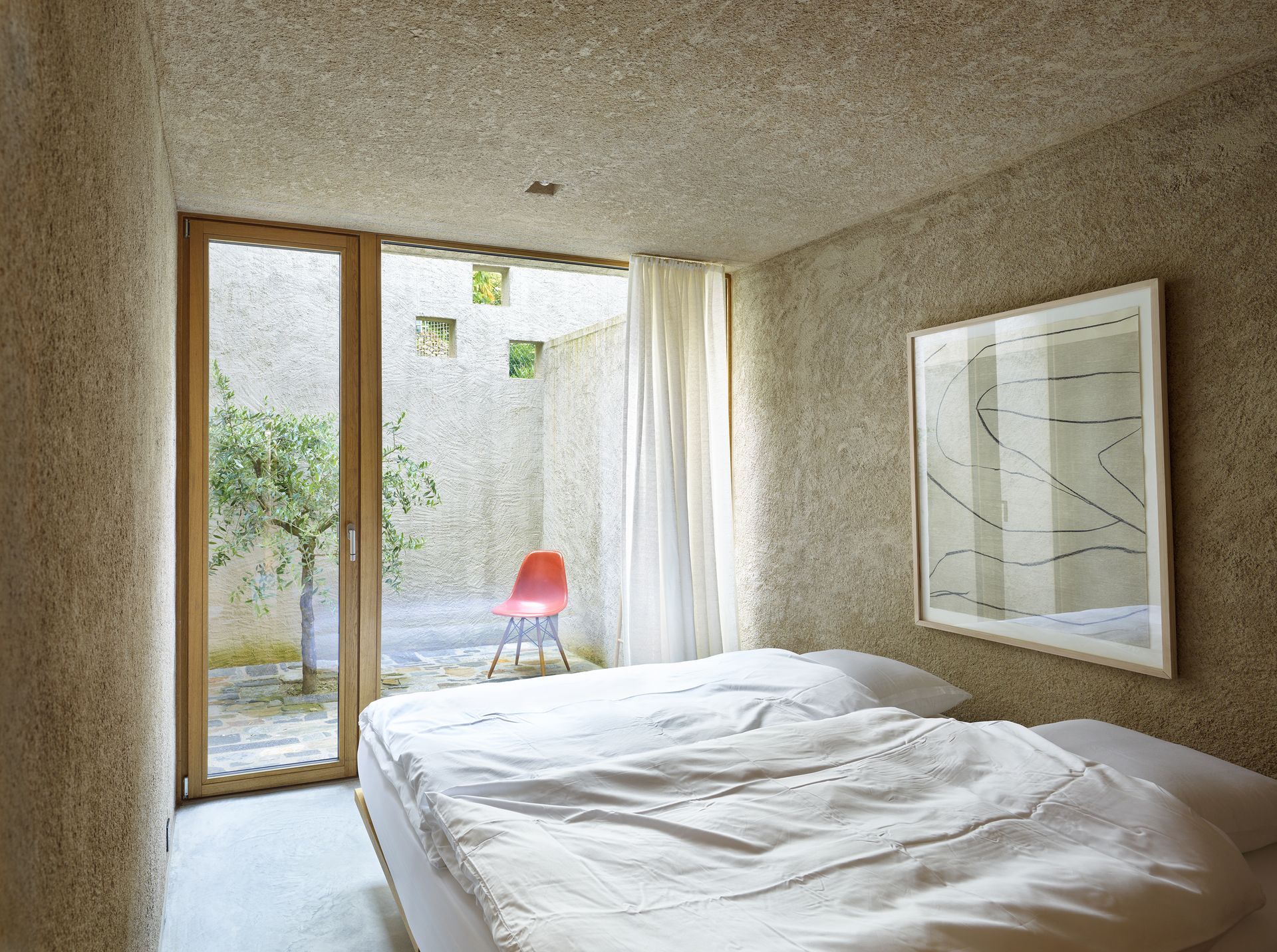 Camera da letto con pareti beige testurizzate, letto, opere d'arte e porta aperta su un piccolo cortile con una sedia rossa.