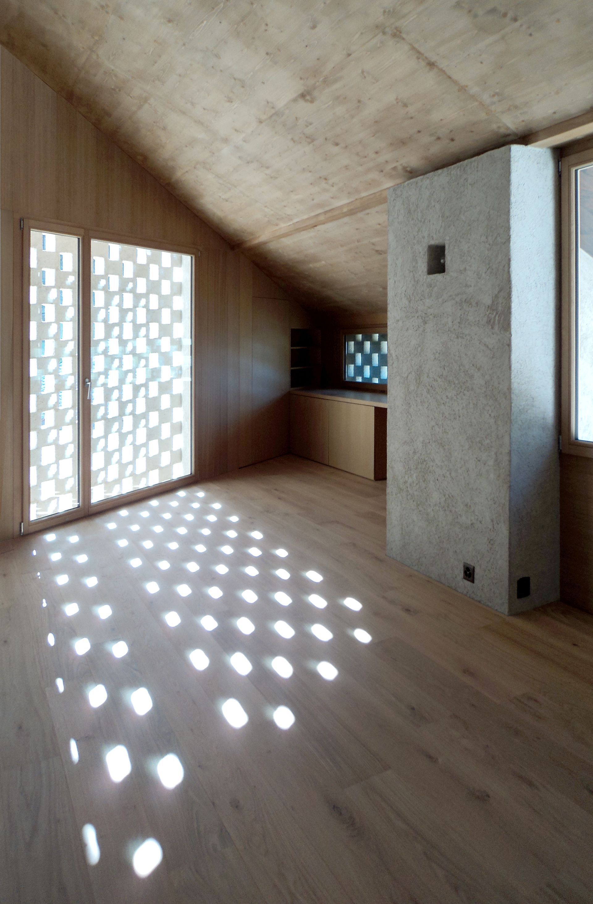 Interno della stanza con pareti in legno e finestre perforate che proiettano motivi di luce solare sul pavimento.