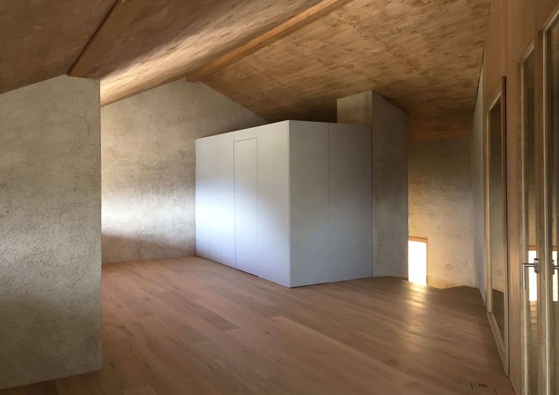 Vista interna di una stanza moderna con pavimenti e soffitto in legno, armadio bianco e pareti dai colori chiari.