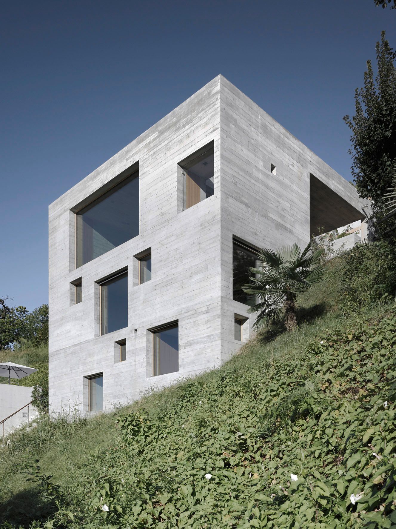 Moderna casa cubica in cemento grigio con finestre di varie dimensioni, costruita sul pendio di una collina con vegetazione.