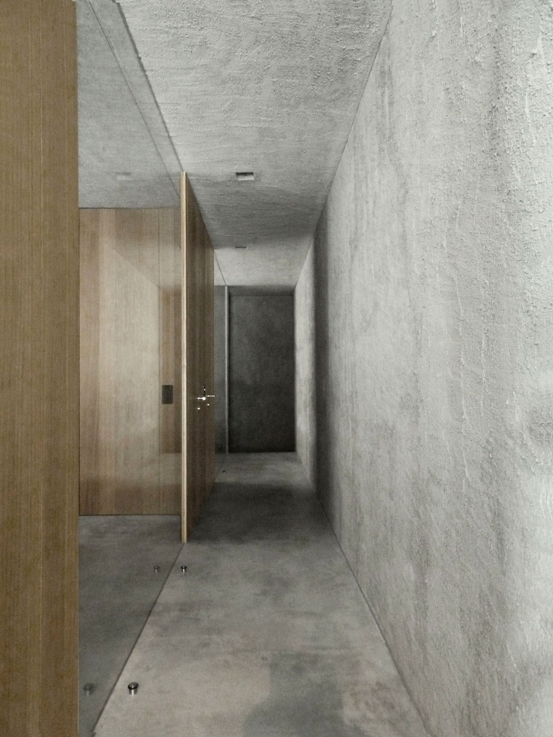 Corridoio stretto con pareti, pavimento e soffitto in cemento. Porte in legno a sinistra e al centro.