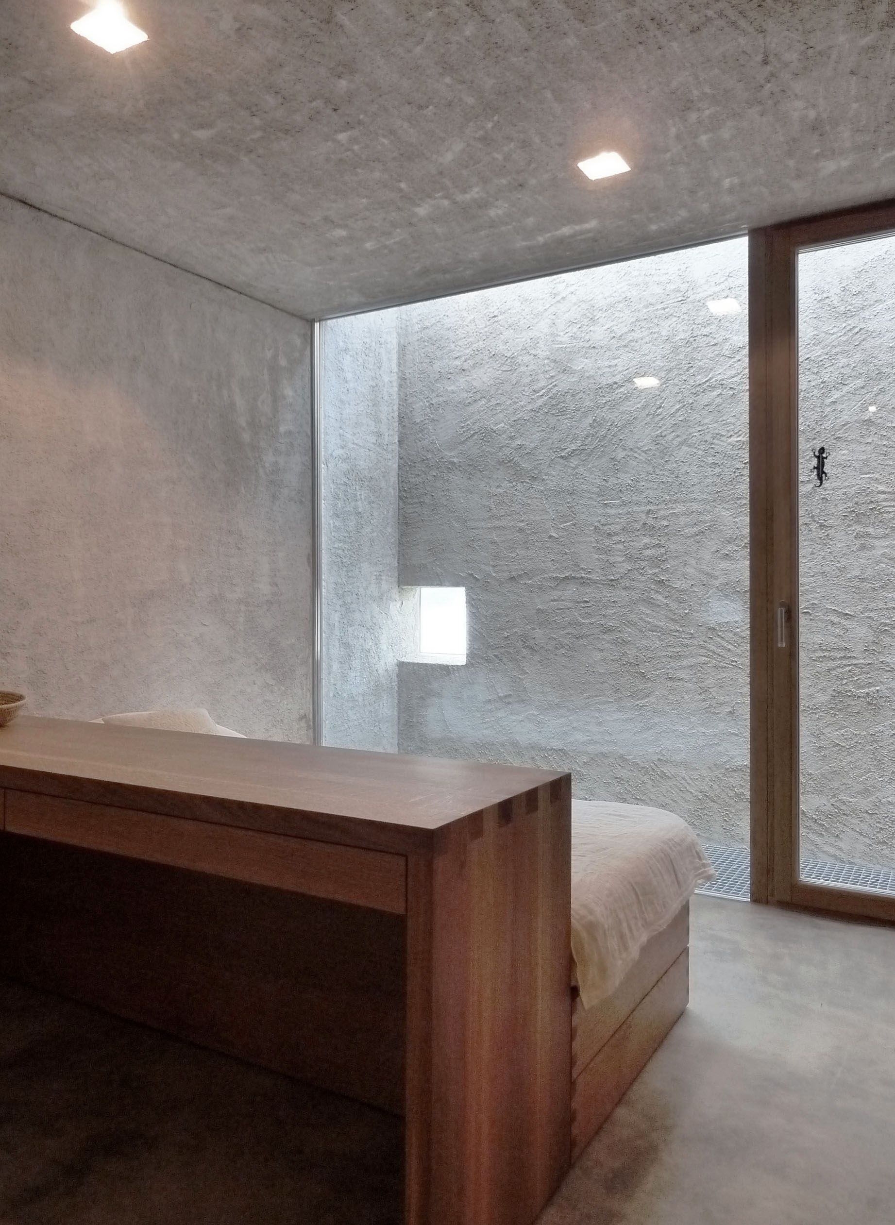 Camera da letto minimalista con scrivania e letto in legno, pareti in cemento e una grande finestra.