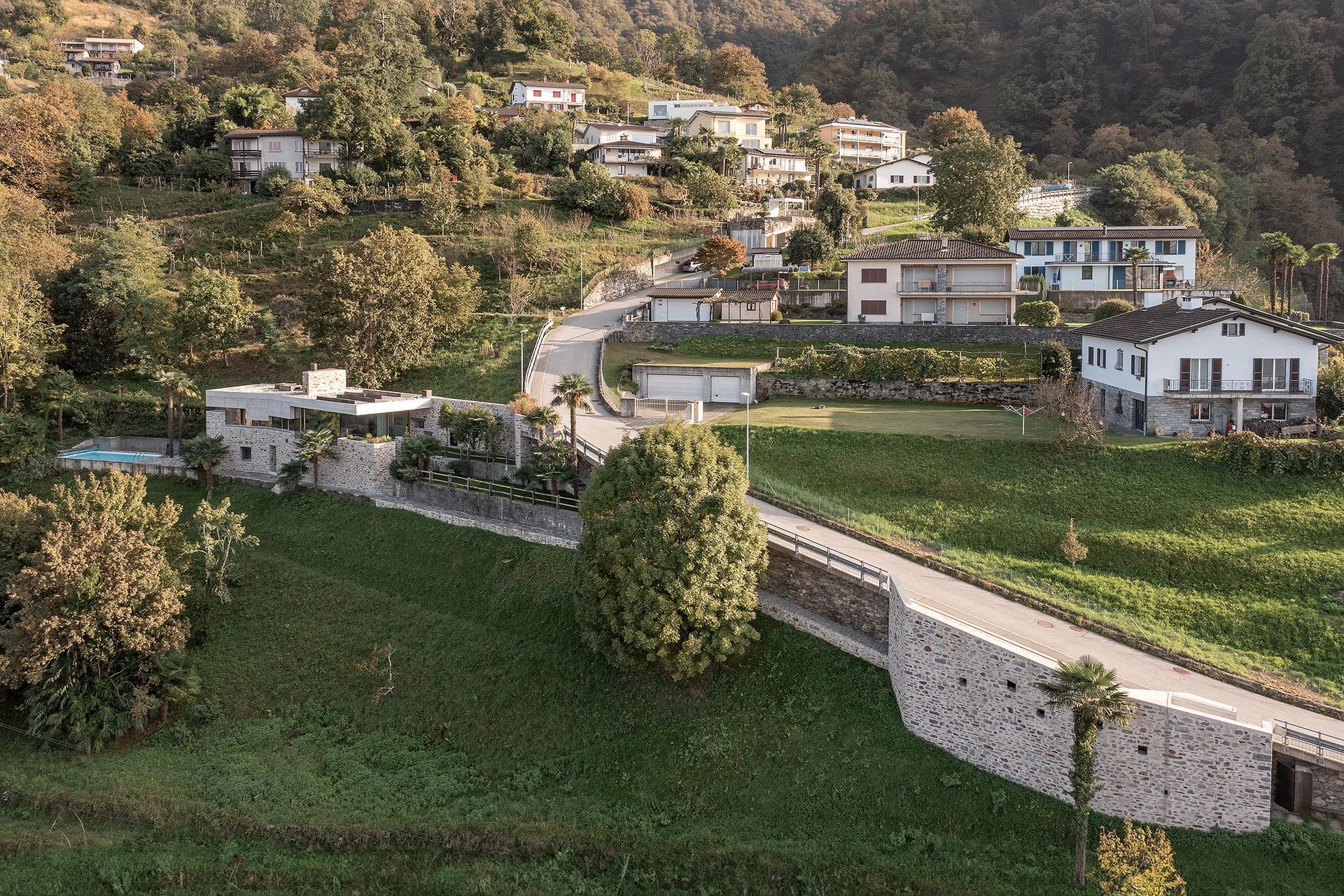 Case su una collina con un muro di pietra e una strada. Erba verde e alberi le circondano.