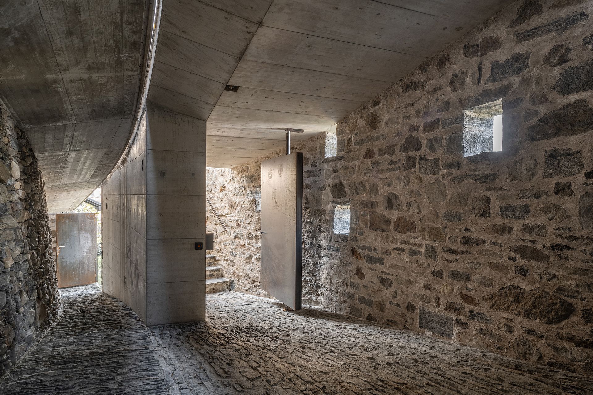 Interno con pareti in pietra, soffitto e porta in cemento, piccole finestre quadrate. Pavimento in ghiaia.
