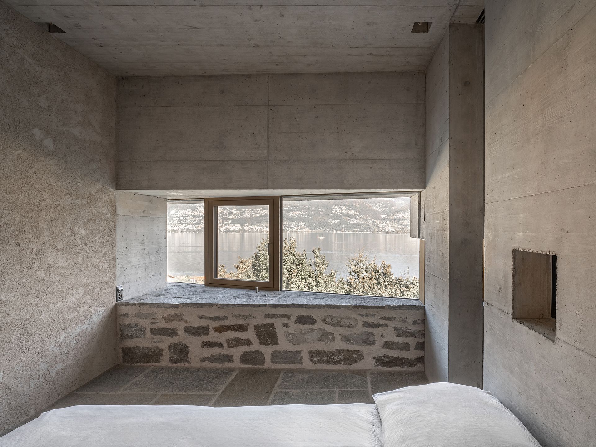 Stanza in cemento con vetrata e muro in pietra che si affaccia sul lago. In primo piano, un letto con un piumone bianco.
